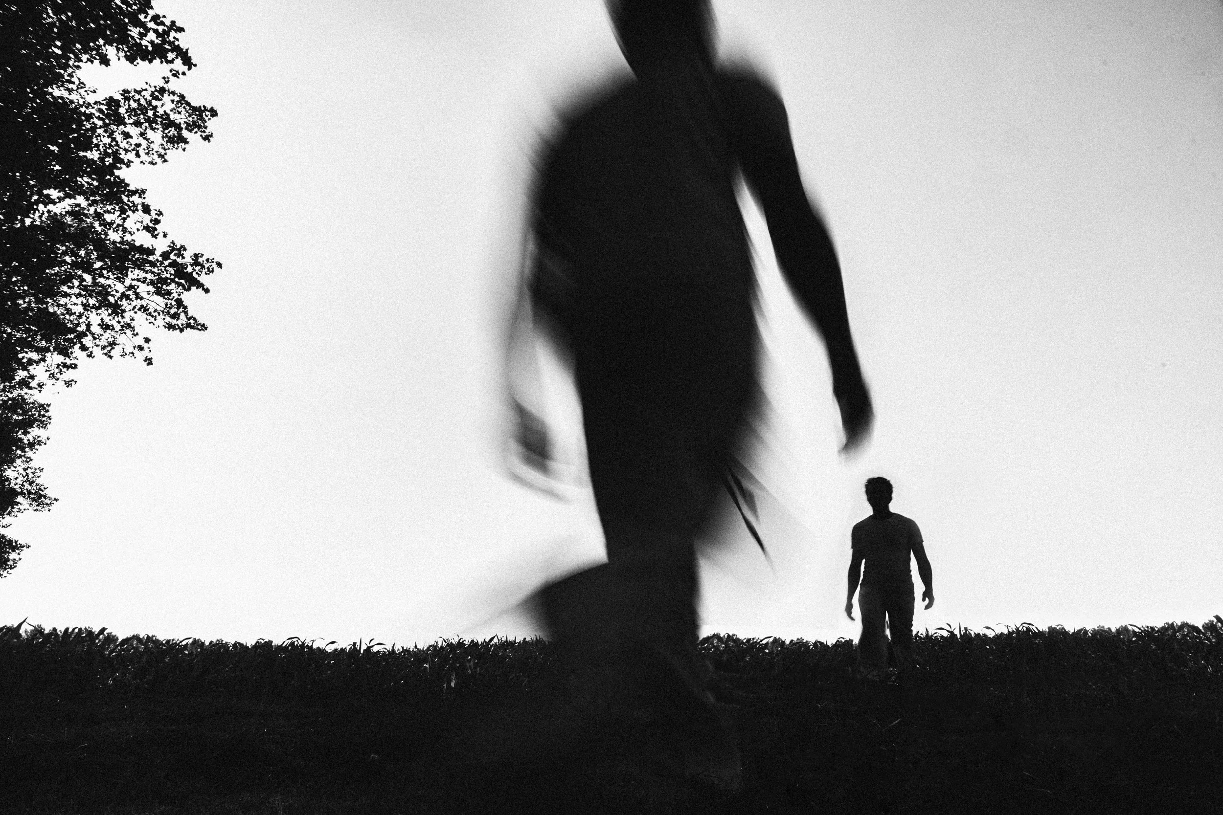 Silhouettes of three people standing outdoors with a tree on the left side and a cloudy sky in the background, captured in black and white.