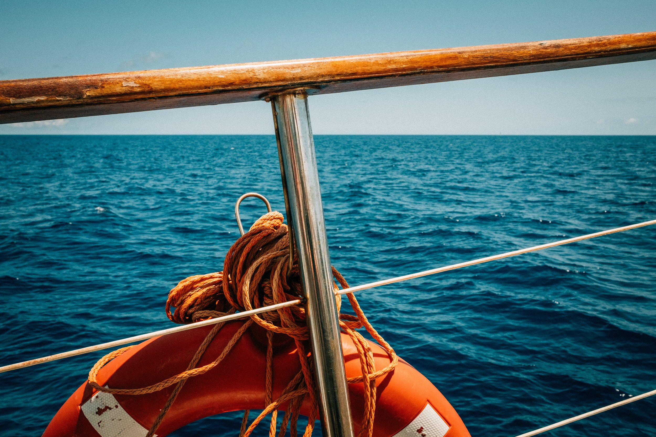 View from a boat showing the ocean, a wooden railing, an orange lifebuoy, and coiled ropes.