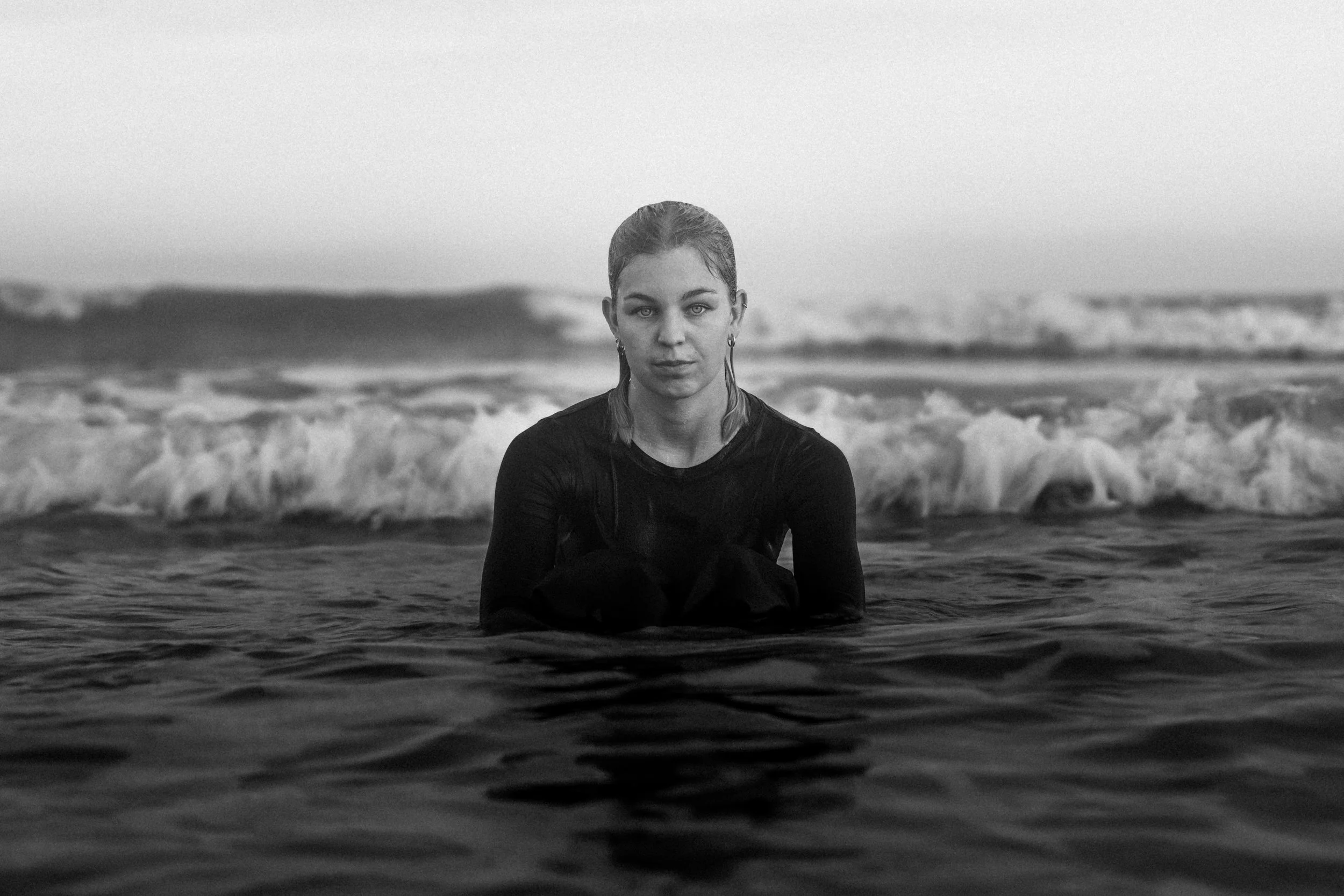 A woman with wet hair sitting in shallow ocean water, with waves crashing behind her and a distant horizon in the background, in black and white.