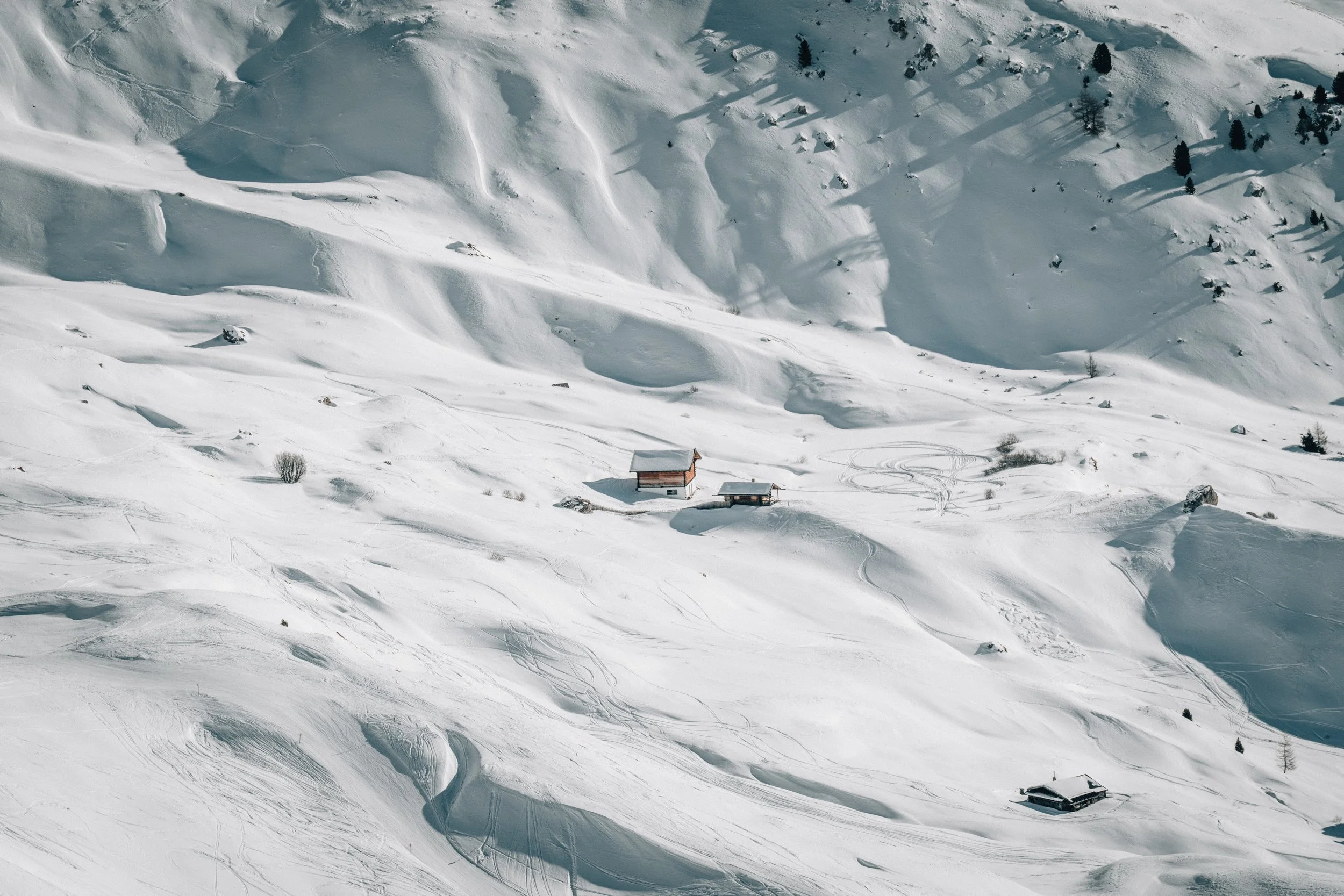 Aerial view of snow-covered mountains with two houses at the base, surrounded by ski tracks and scattered trees.