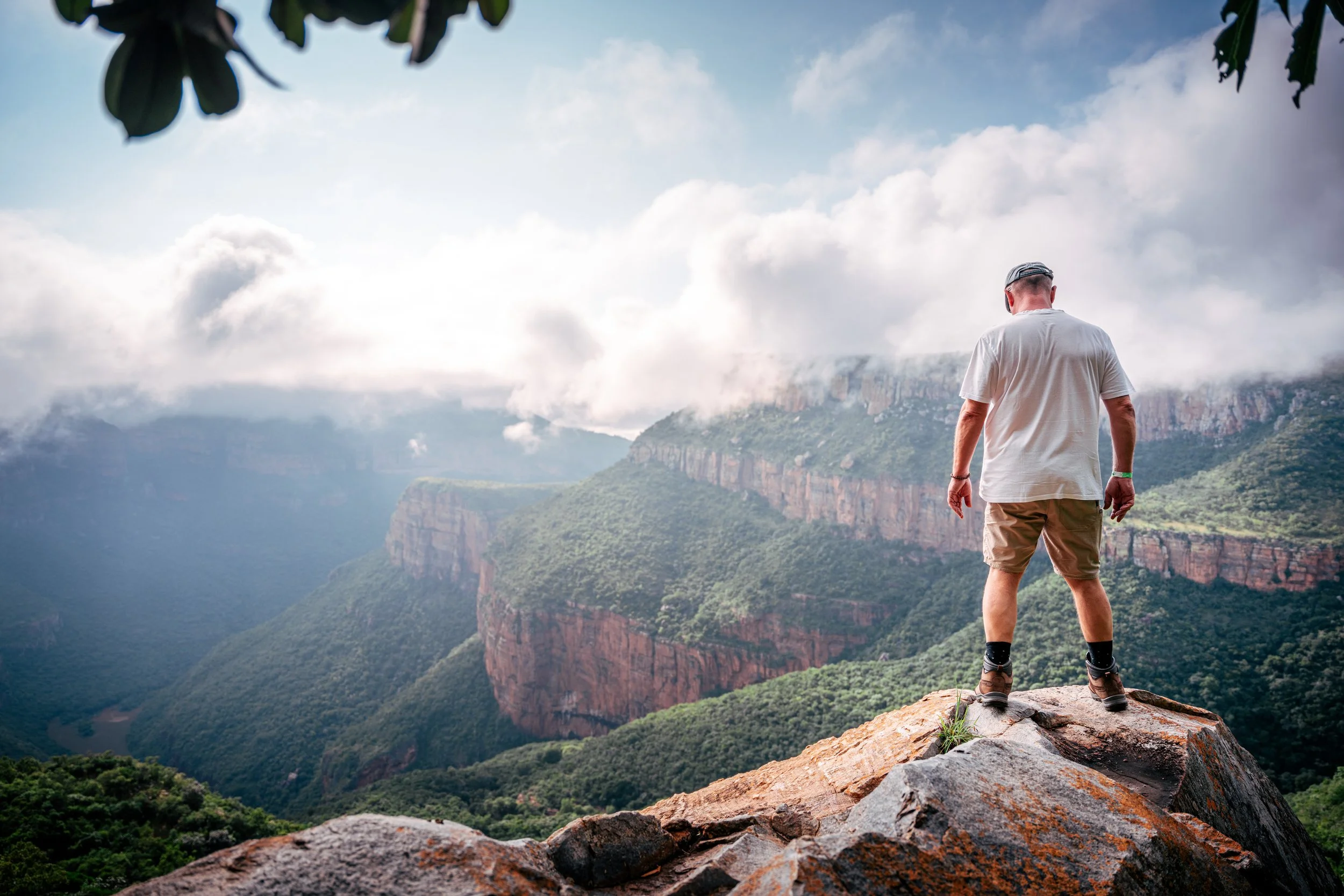A man standing on the edge of a rocky cliff overlooking a lush green canyon with layers of cliffs and mountains in the distance, under a partly cloudy sky.