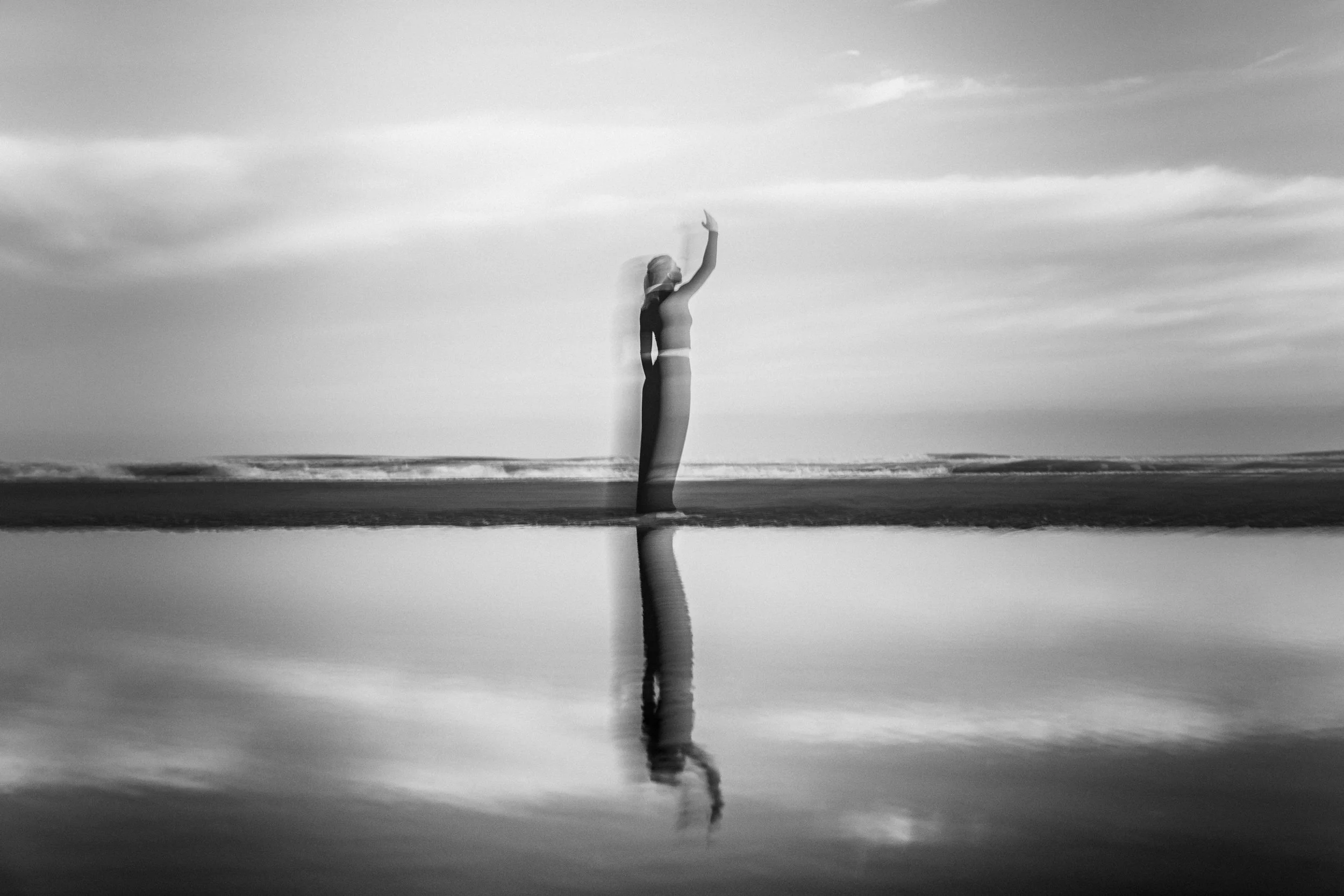 Black and white photograph of a woman in a dress standing on a beach, raising one arm, with her reflection visible in the wet sand, and clouds in the sky.