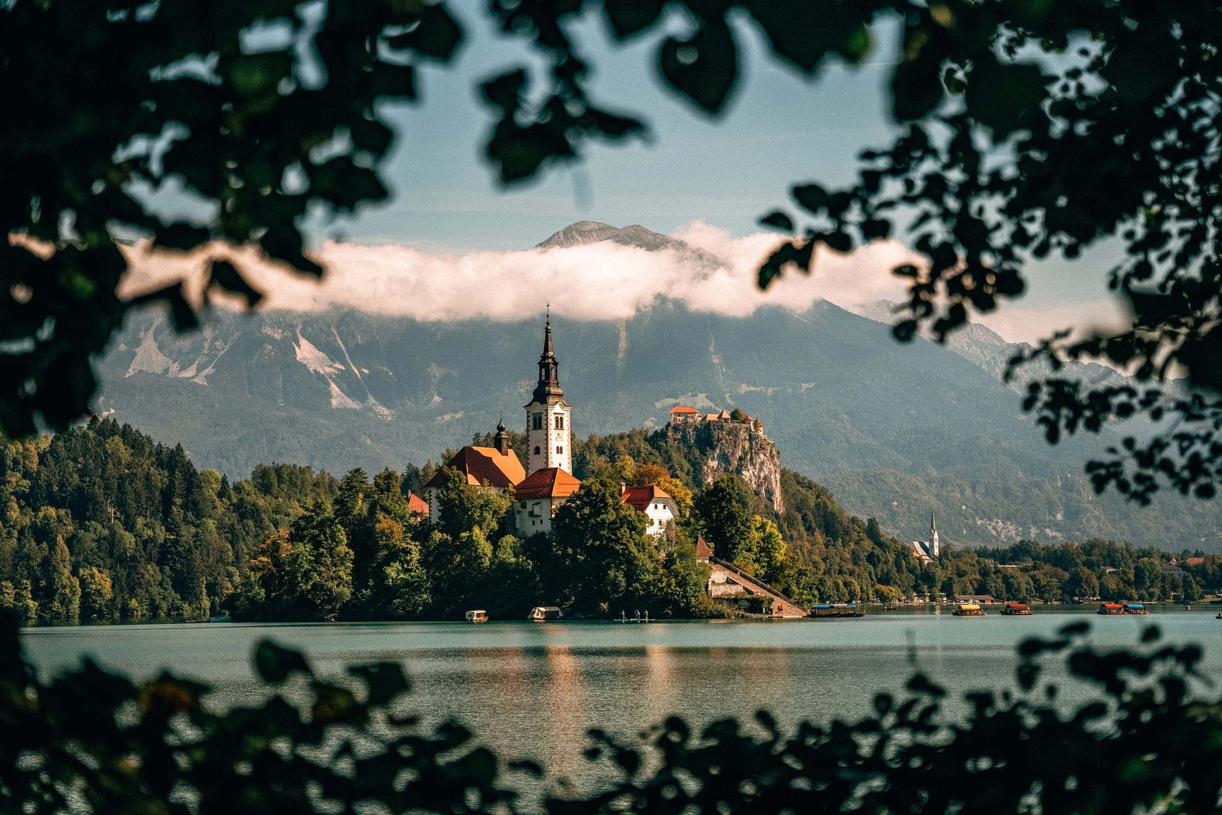 A scenic view of a lakeside village with a church and other buildings atop a hill, surrounded by lush green trees, with mountain peaks and clouds in the background, framed by dark leaves in the foreground.