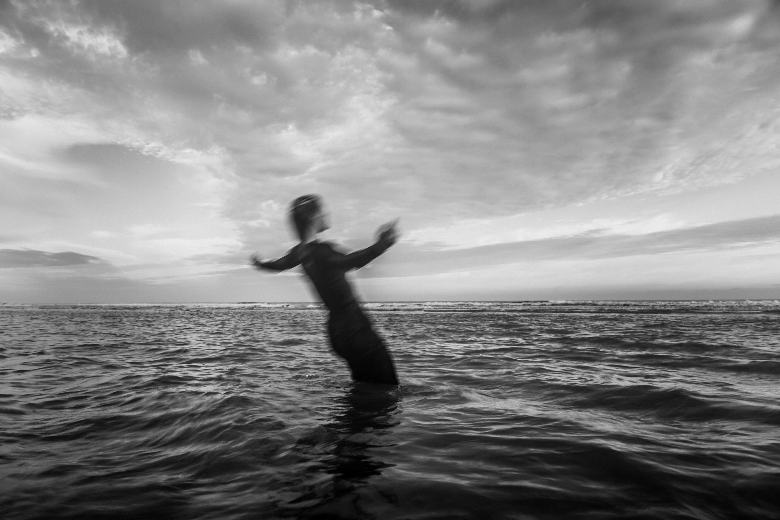 A person in the ocean appears to be flipping or falling into the water, with a cloudy sky above.
