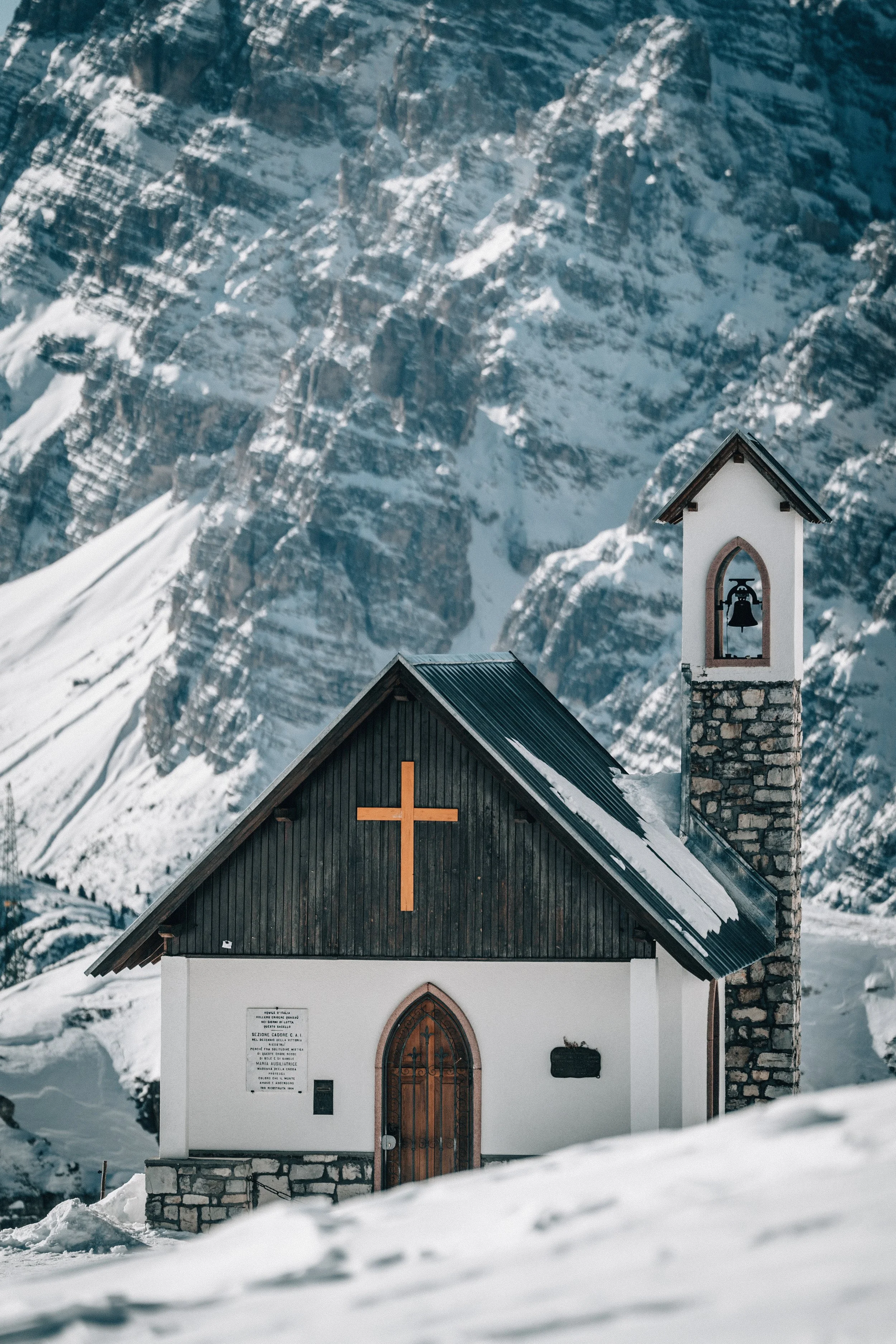A small church with a wooden cross on the front facade, situated in a snow-covered landscape with a mountainous backdrop. The church has a white exterior, a wooden door, and a bell tower with a bell visible inside.