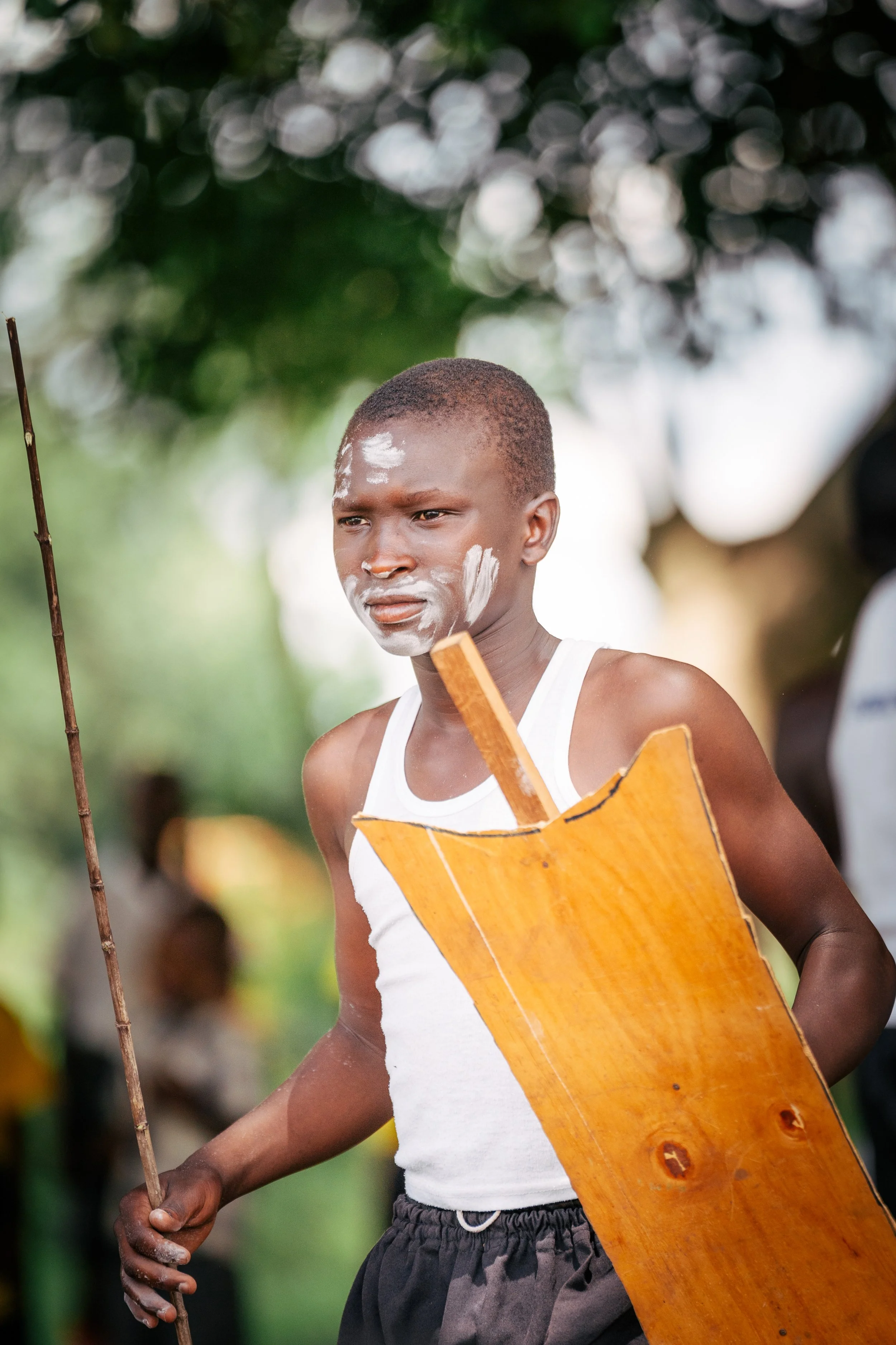 A young boy with face paint on his cheeks and forehead, wearing a white tank top and dark shorts, holding a wooden shield and stick, outdoors with blurred greenery in the background.