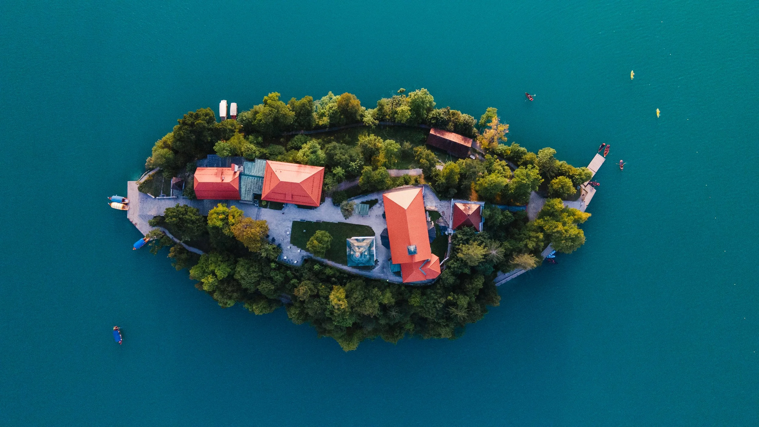 Aerial view of a small island with trees, buildings with red roofs, a dock, and boats on surrounding blue water.
