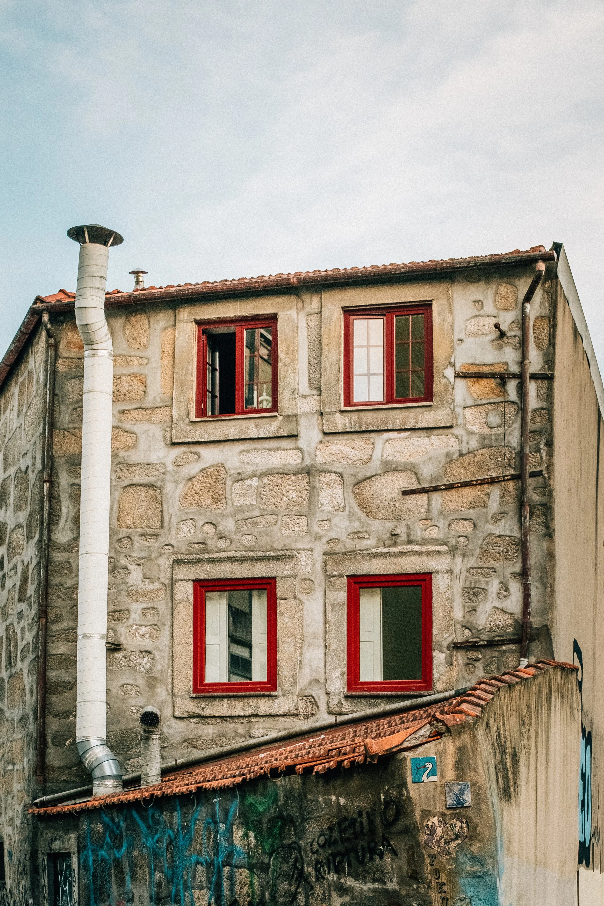 A stone building with red-framed windows, a tiled roof, and graffiti on the lower section. A white vent pipe extends from the top left corner. The sky is cloudy.
