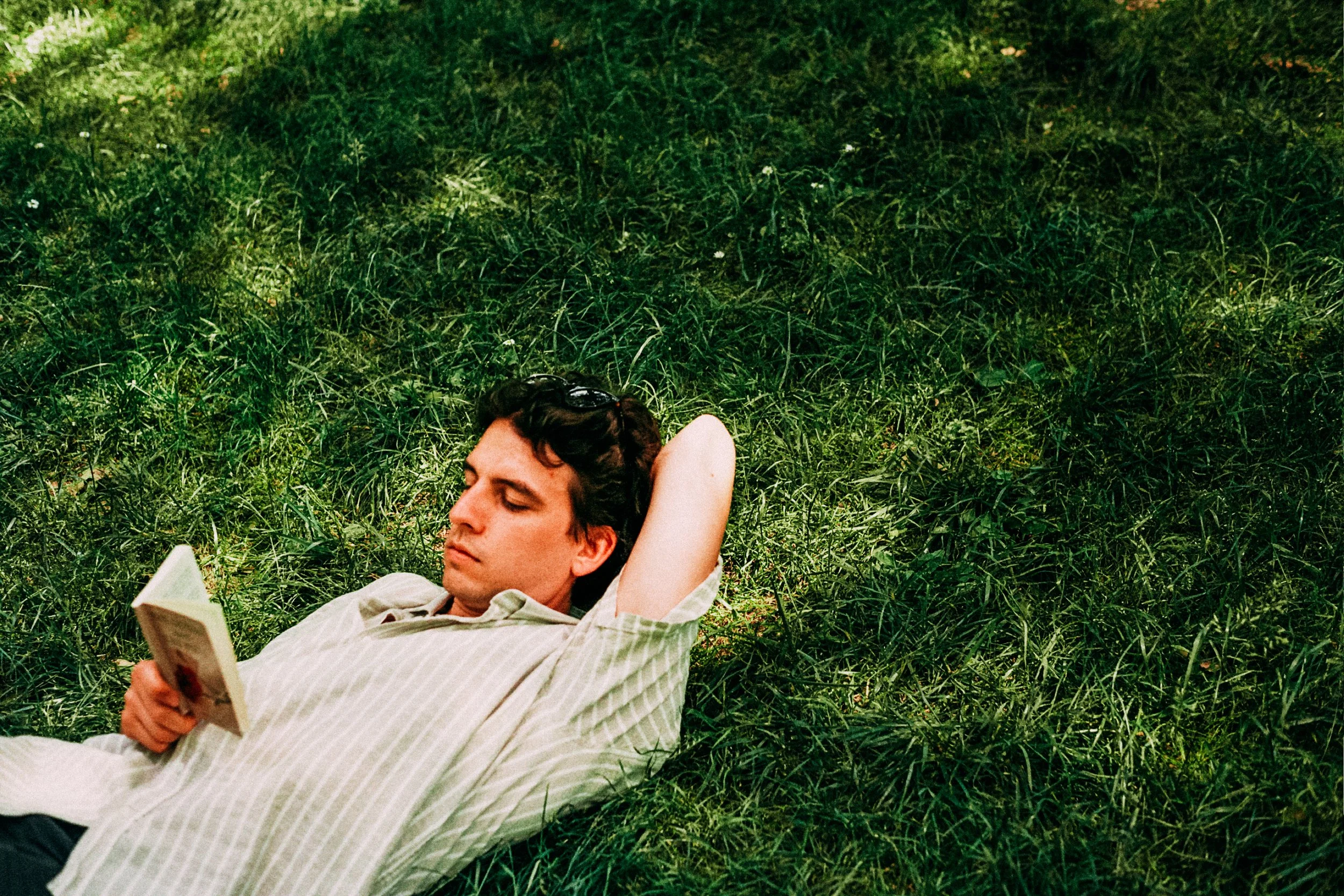A young man with curly hair is lying on green grass, reading a book, with sunglasses resting on his forehead and one arm behind his head.