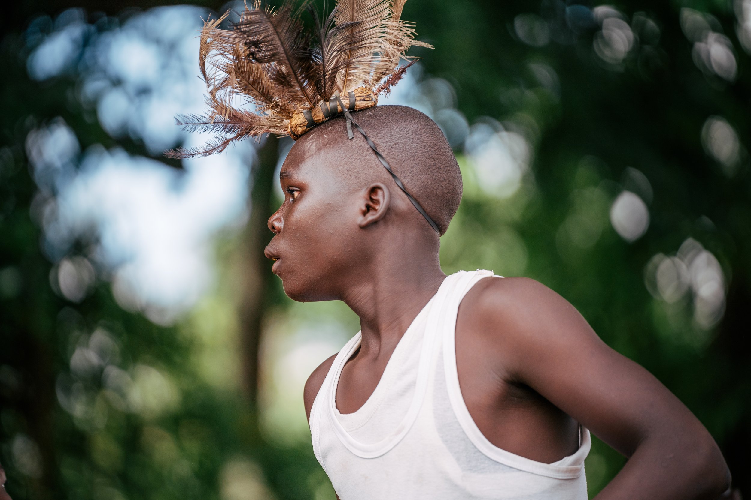 A person with a shaved head and dark skin wearing a white tank top, adorned with a headdress made of feathers, outdoors with a blurred background of green trees.