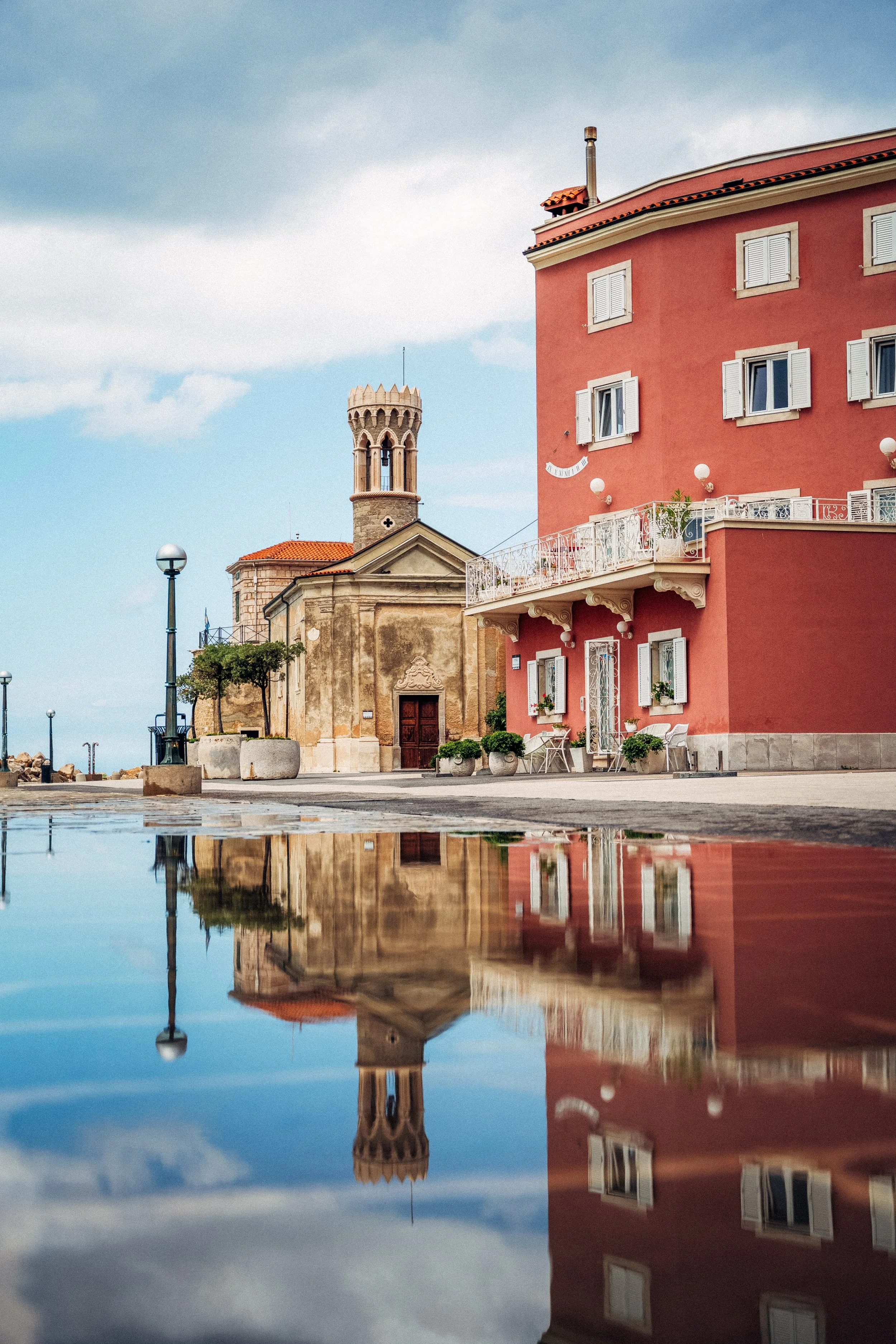 A reflections of buildings in a puddle of water on the ground, showing a red multi-story building, a historic tower, and part of another older building with a cross on its roof, with cloudy sky overhead.