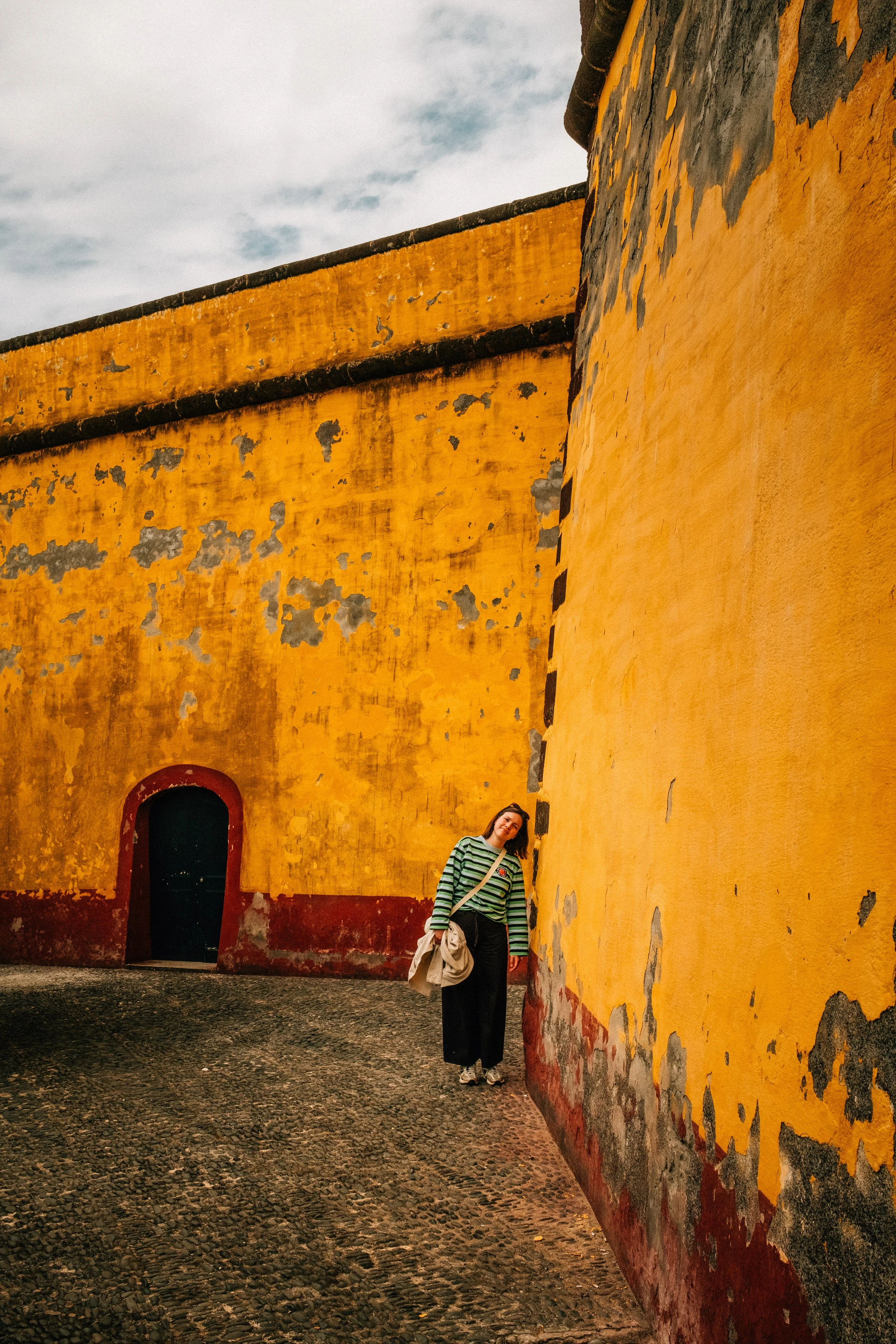 A woman stands in front of an old, weathered yellow wall with peeling paint and a small arched doorway. She is wearing a green and black striped sweater, black pants, and sneakers, holding her jacket and a bag, with a slight smile. The cobblestone gr