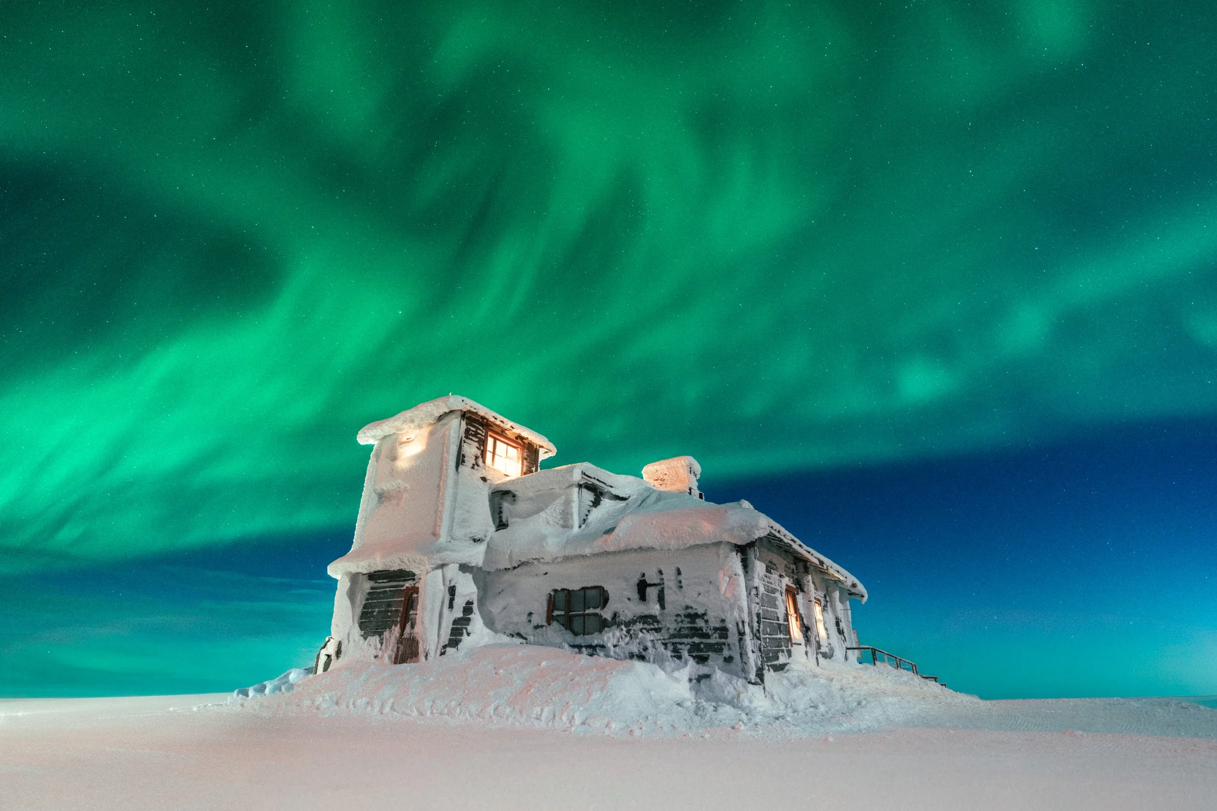 A snow-covered house at night under the northern lights, with a dark sky and visible stars.