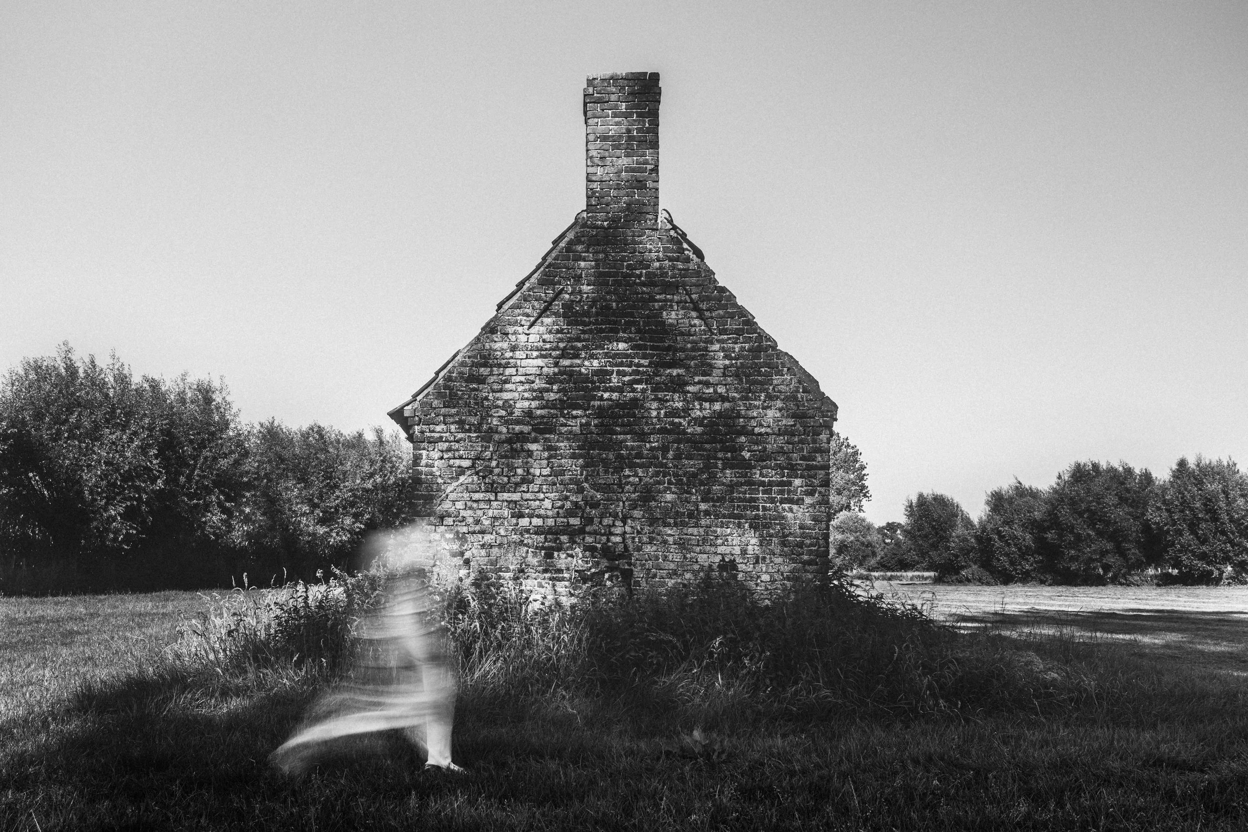 Black and white photo of an old brick house with a chimney, surrounded by trees and grass, with a blurred figure of a dog in the foreground.