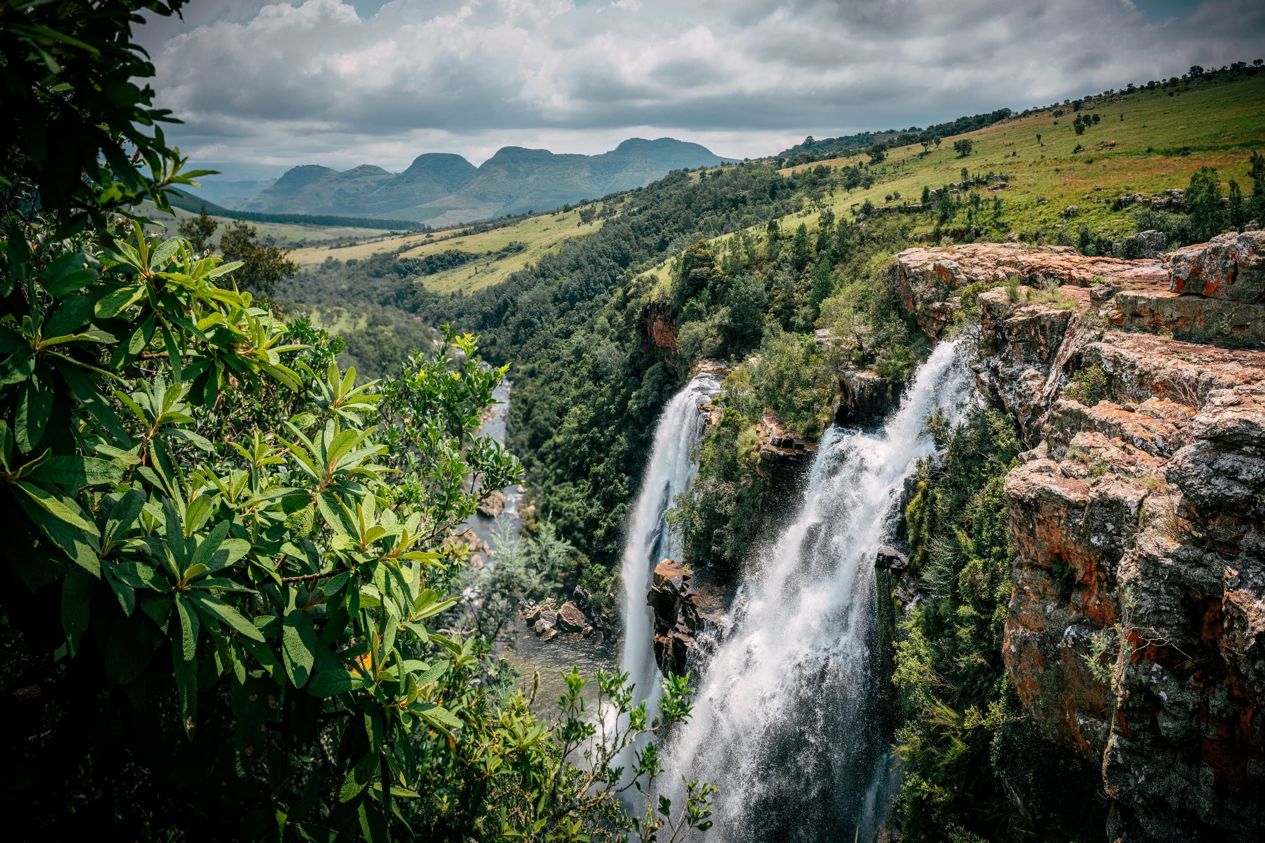 A scenic view of a waterfall cascading down rocks in a lush green valley with mountains in the distance and cloudy skies above.