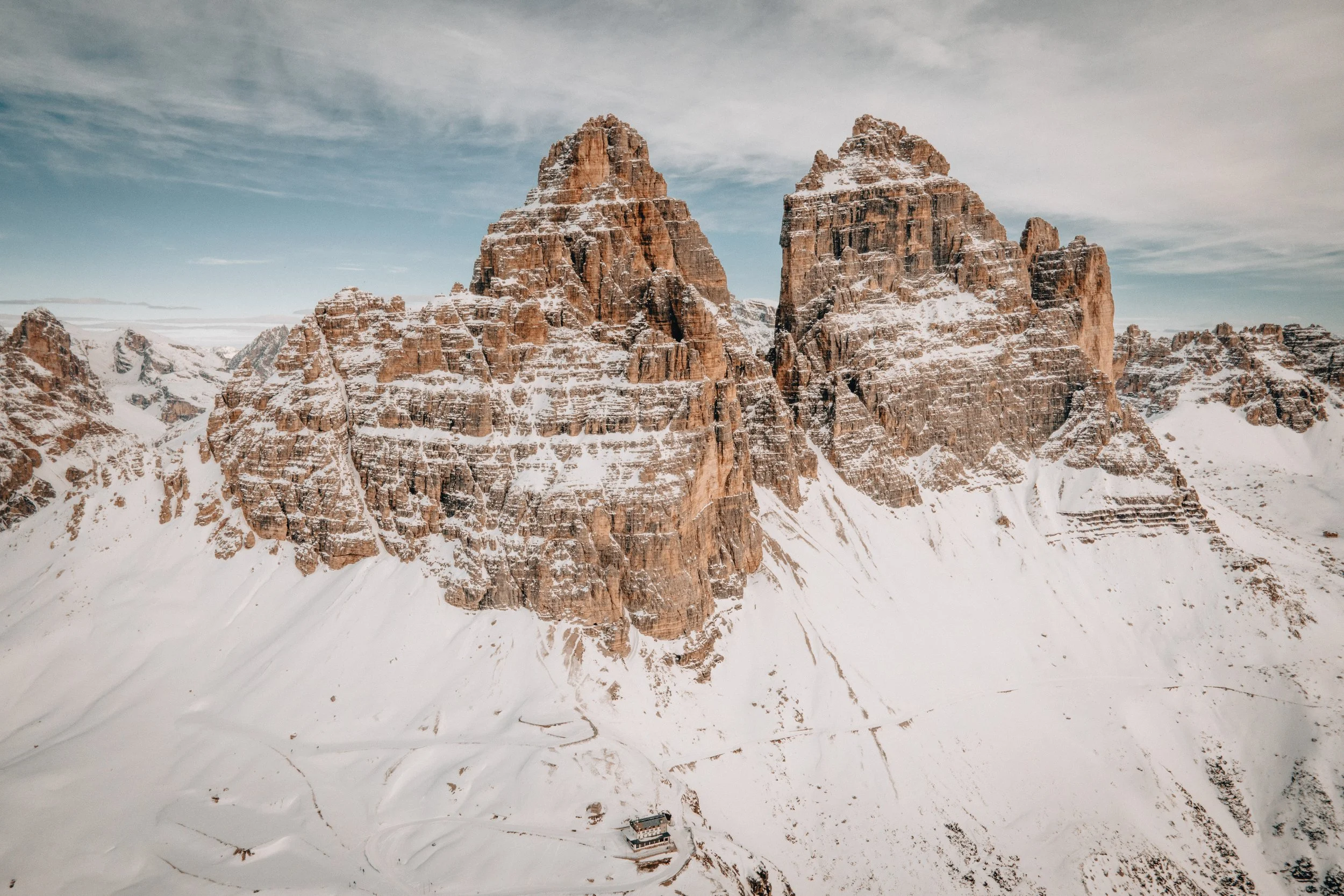 Snow-covered mountain peaks with rugged, reddish-brown rock formations, a cloudy sky, and a small building or structure at the base of the mountains.