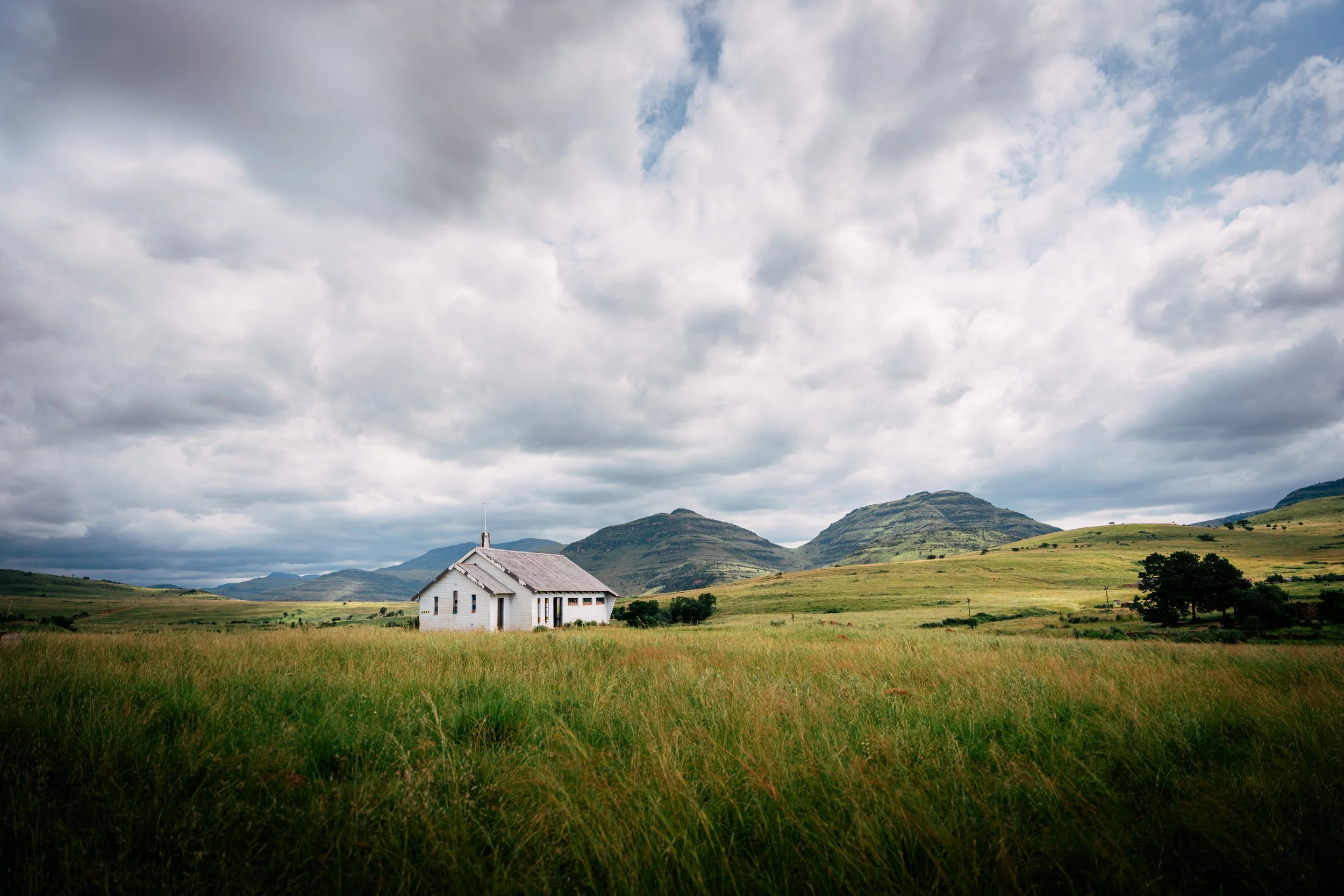 A white church with a steeple surrounded by green grass and rolling hills under a partly cloudy sky.