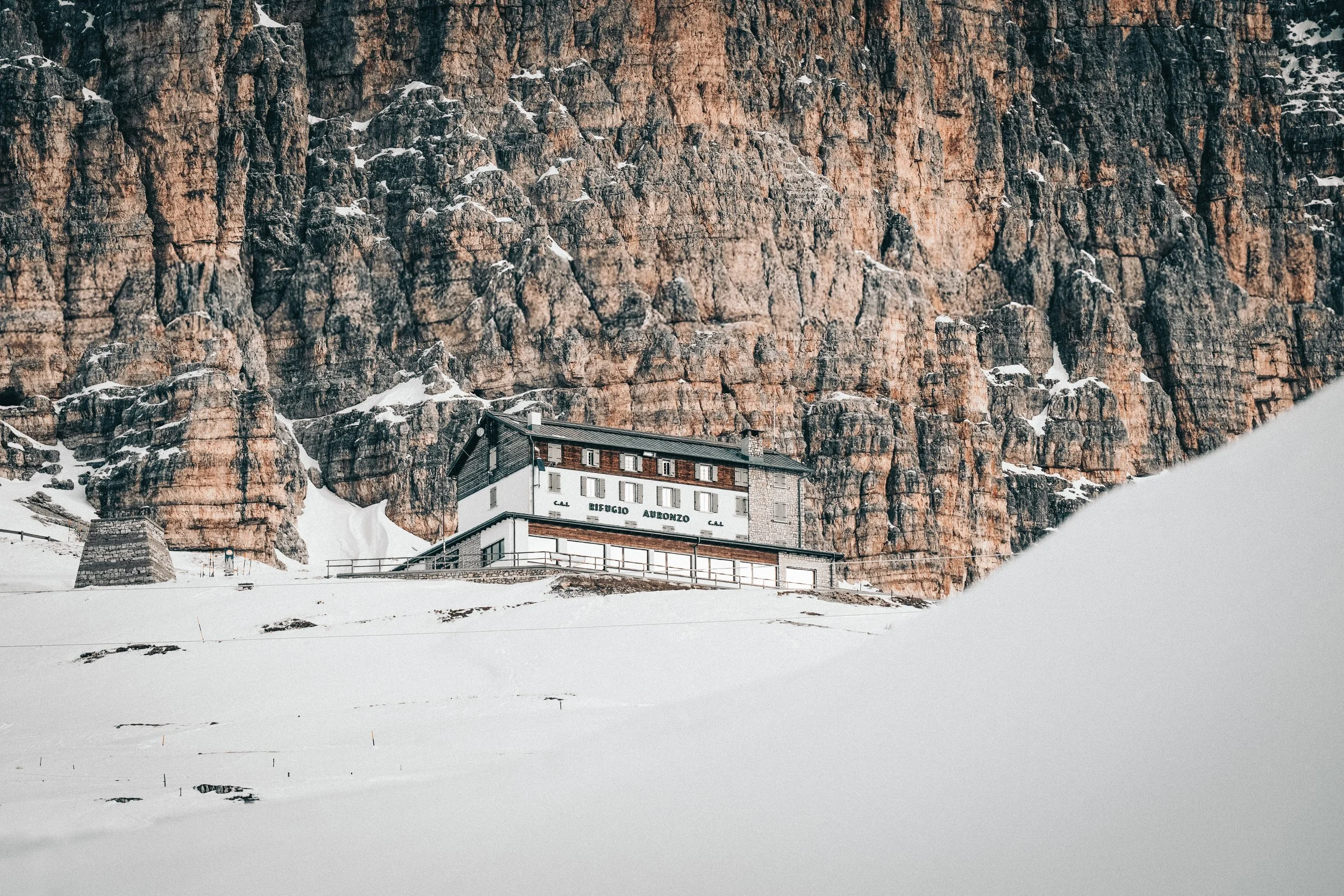 A mountain refuge building called 'Rifugio Auronzo' is situated at the base of a steep rocky mountain face. The building is white with dark trim and has multiple stories. The area around the refuge is covered in snow, and the rocky mountain face rise