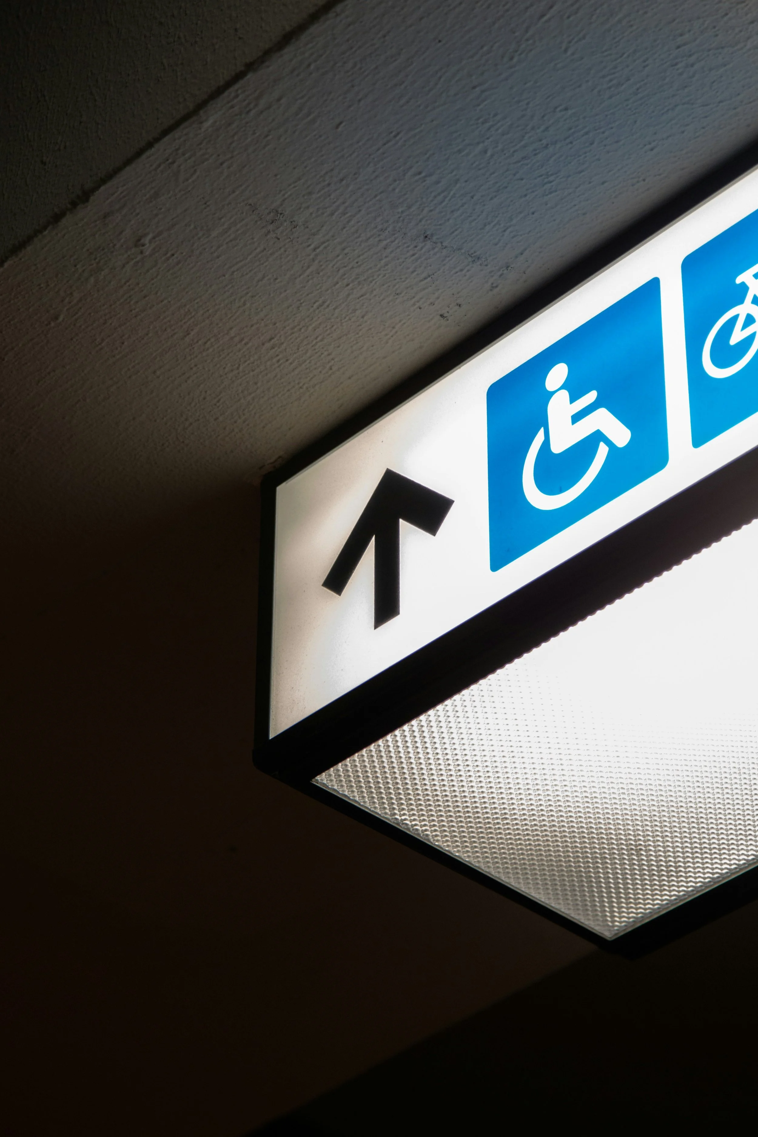 Indoor sign with upward arrow, indicating directions for accessible facilities for disabled individuals and bicycles, mounted on a ceiling with textured surface.