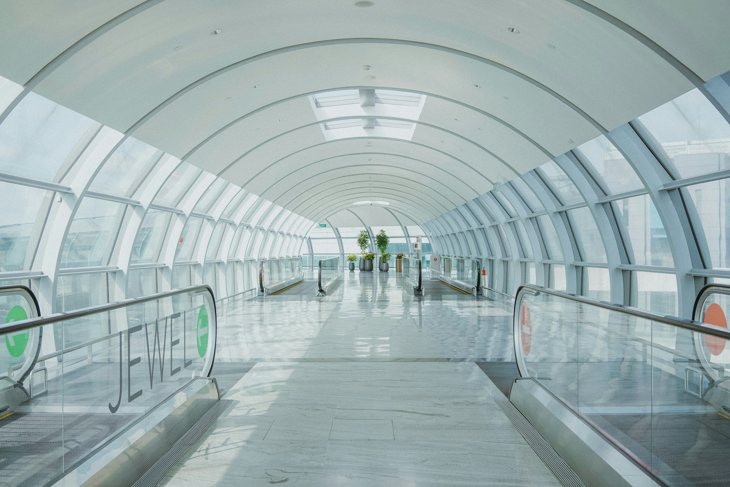 Empty enclosed walkway with glass walls and ceiling, plants in pots at the end, and a moving walkway.