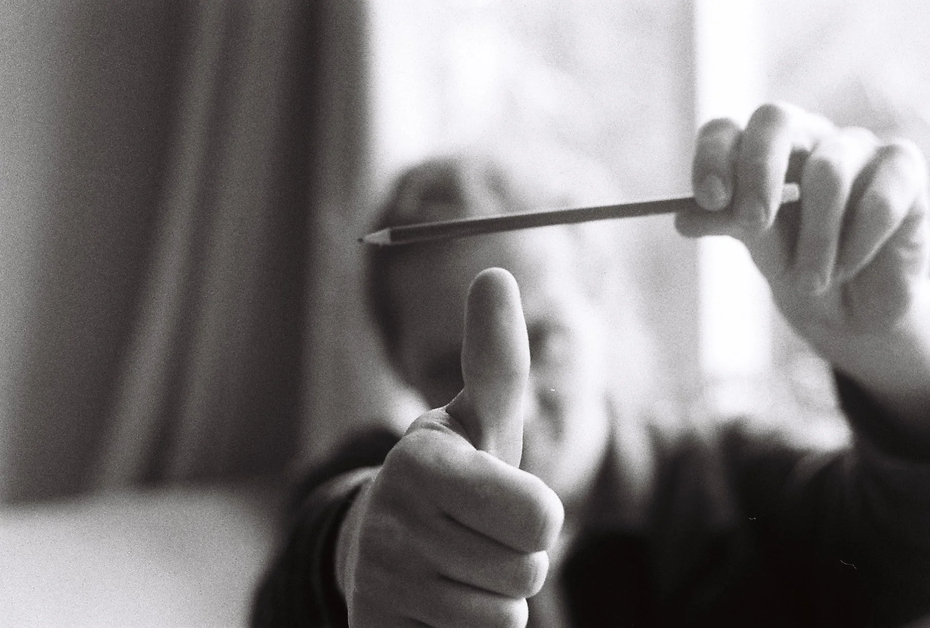A person holding and pointing a pencil towards the camera while giving a thumbs-up, black and white photo with a blurred background.