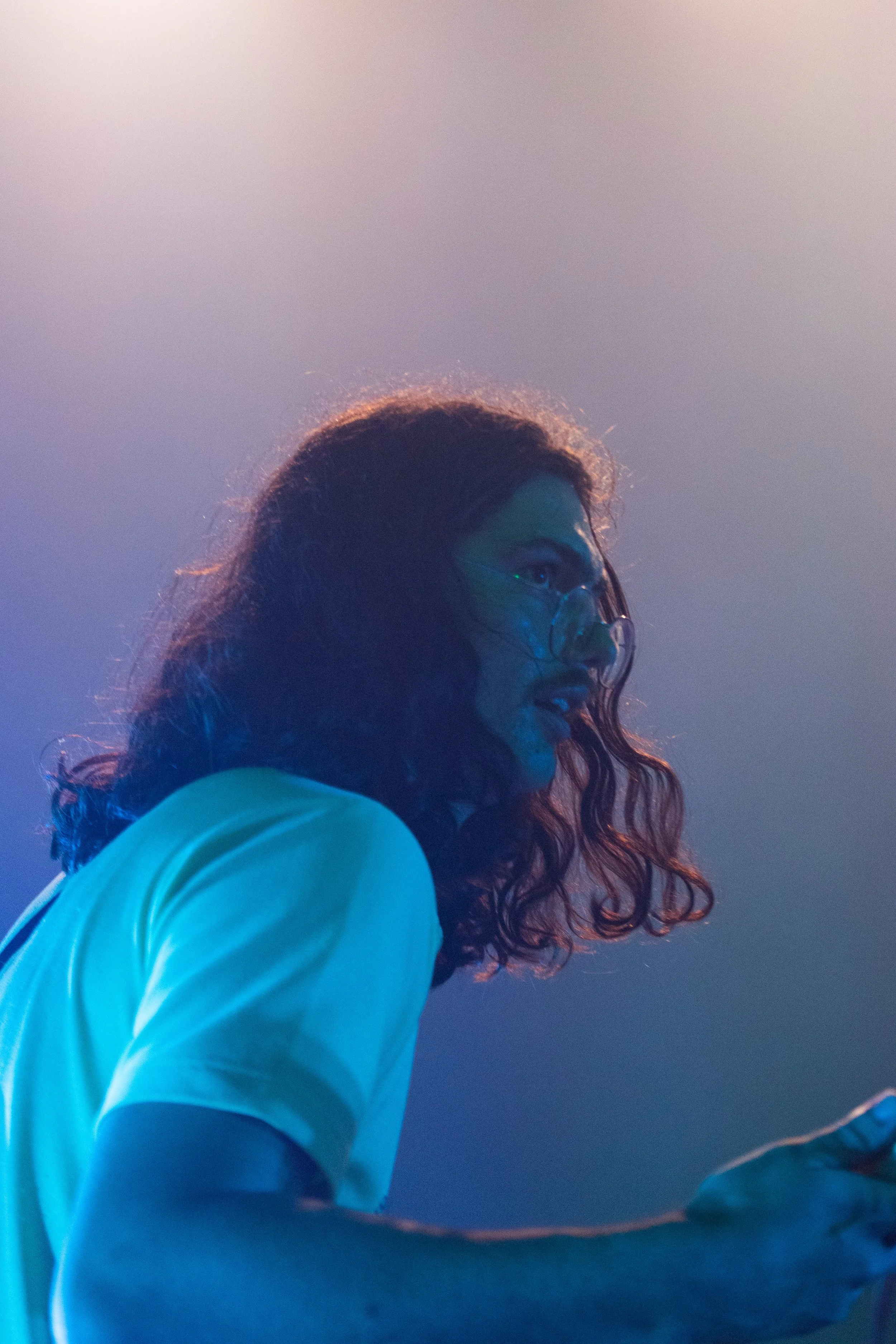 Side profile of a woman with curly hair and glasses, wearing a light blue shirt, against a gradient background of soft pastel colors.