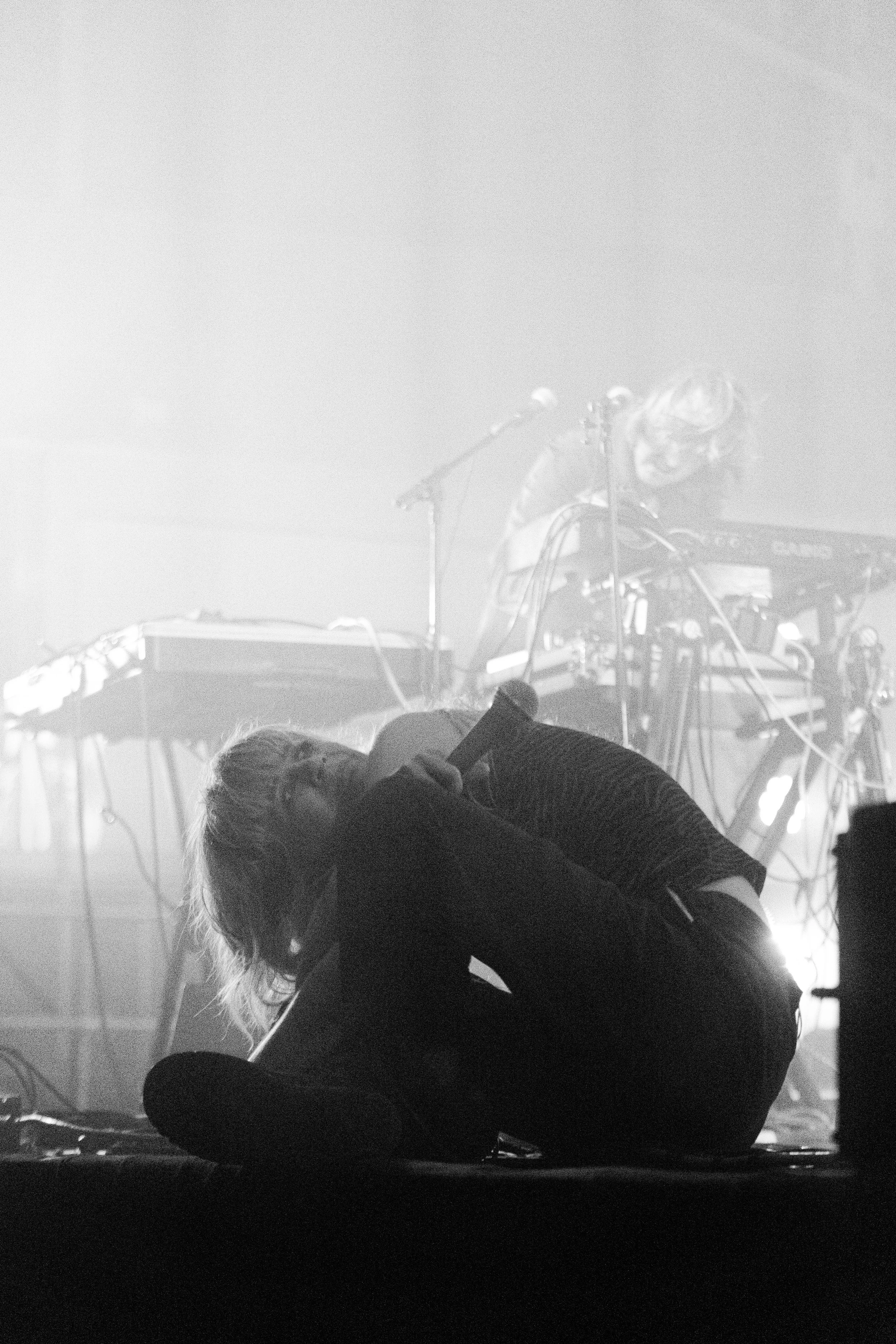 A person with light-colored hair is performing on stage, leaning forward with their head down and one arm resting on their knee. The stage setup includes musical instruments and equipment, with bright lights and fog creating a dramatic atmosphere.