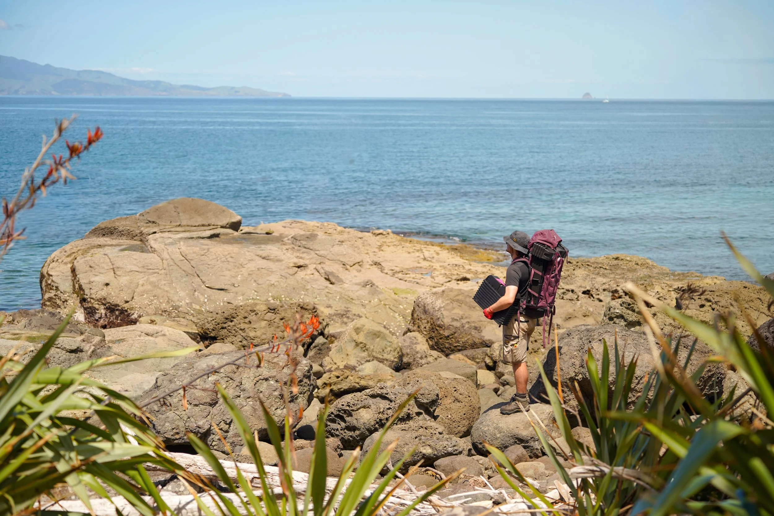 A hiker with a backpack and sun hat standing on rocks by the ocean, with a person carrying a large backpack and walking along the rocky shoreline, overlooking the water and distant landforms.