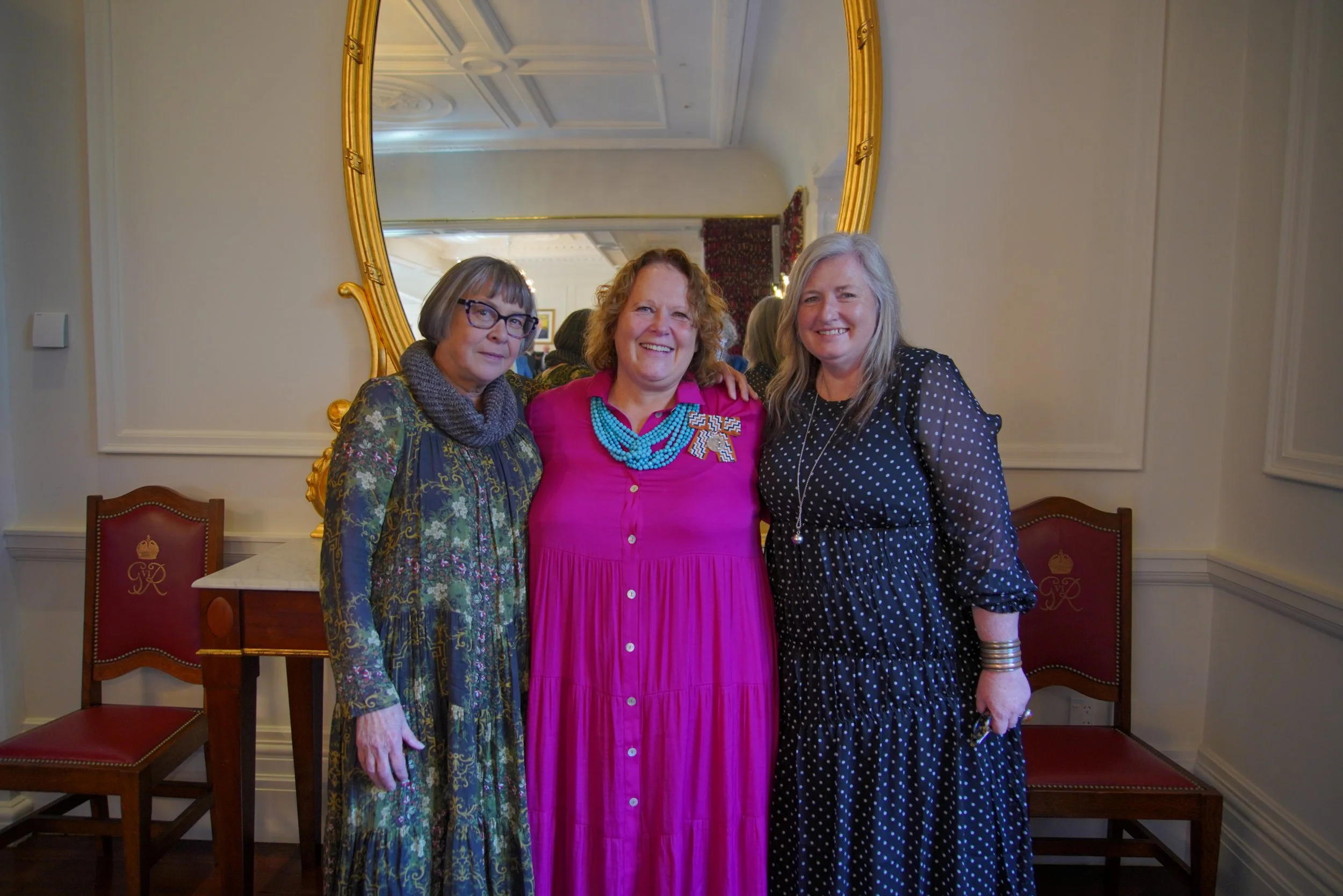 Three women standing close together in a room with a large ornate gold mirror behind them. They are smiling and dressed in colorful clothing, with two of them wearing glasses.