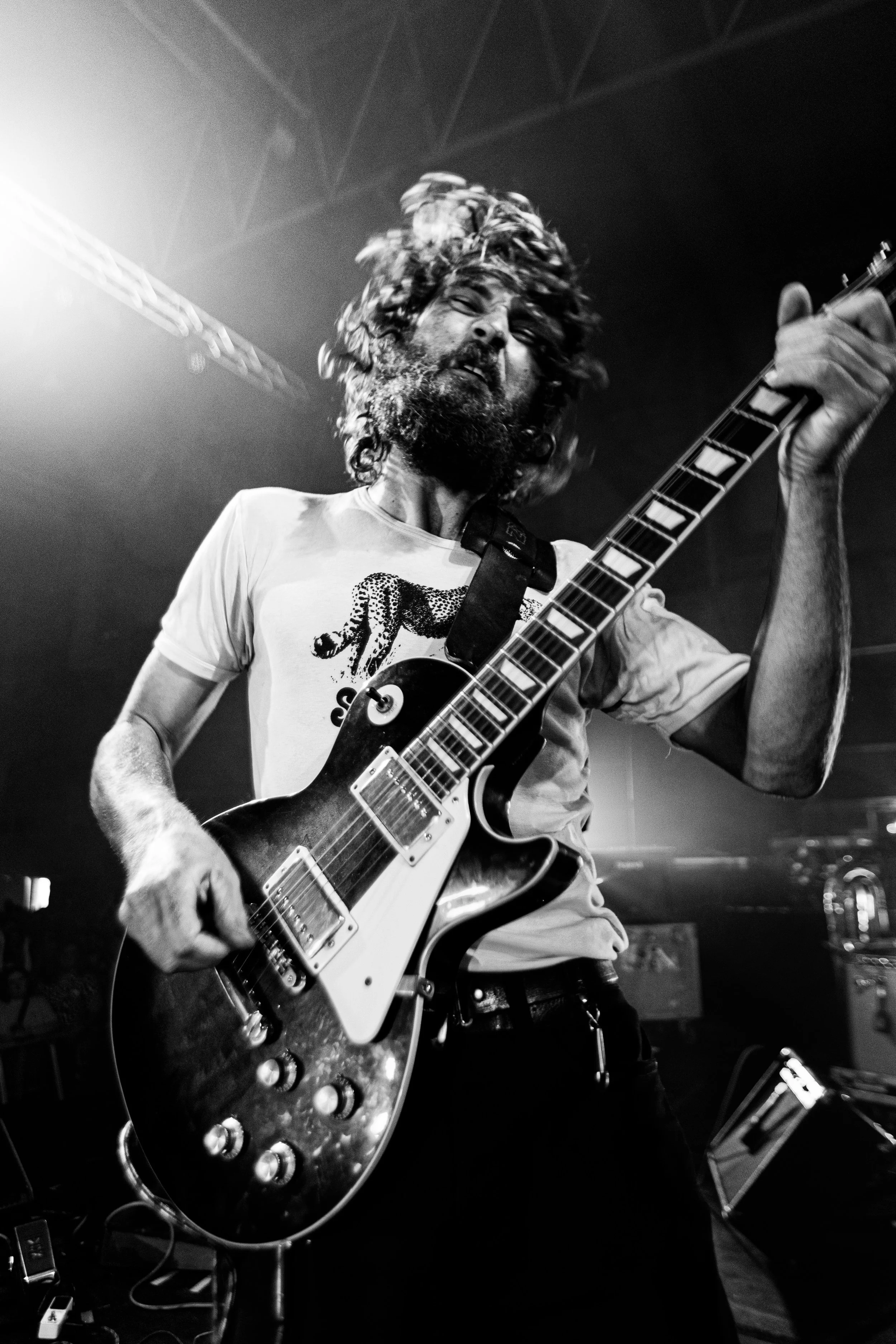 A man with long curly hair and a beard passionately playing an electric guitar on stage in a black and white photo, with stage lighting in the background.