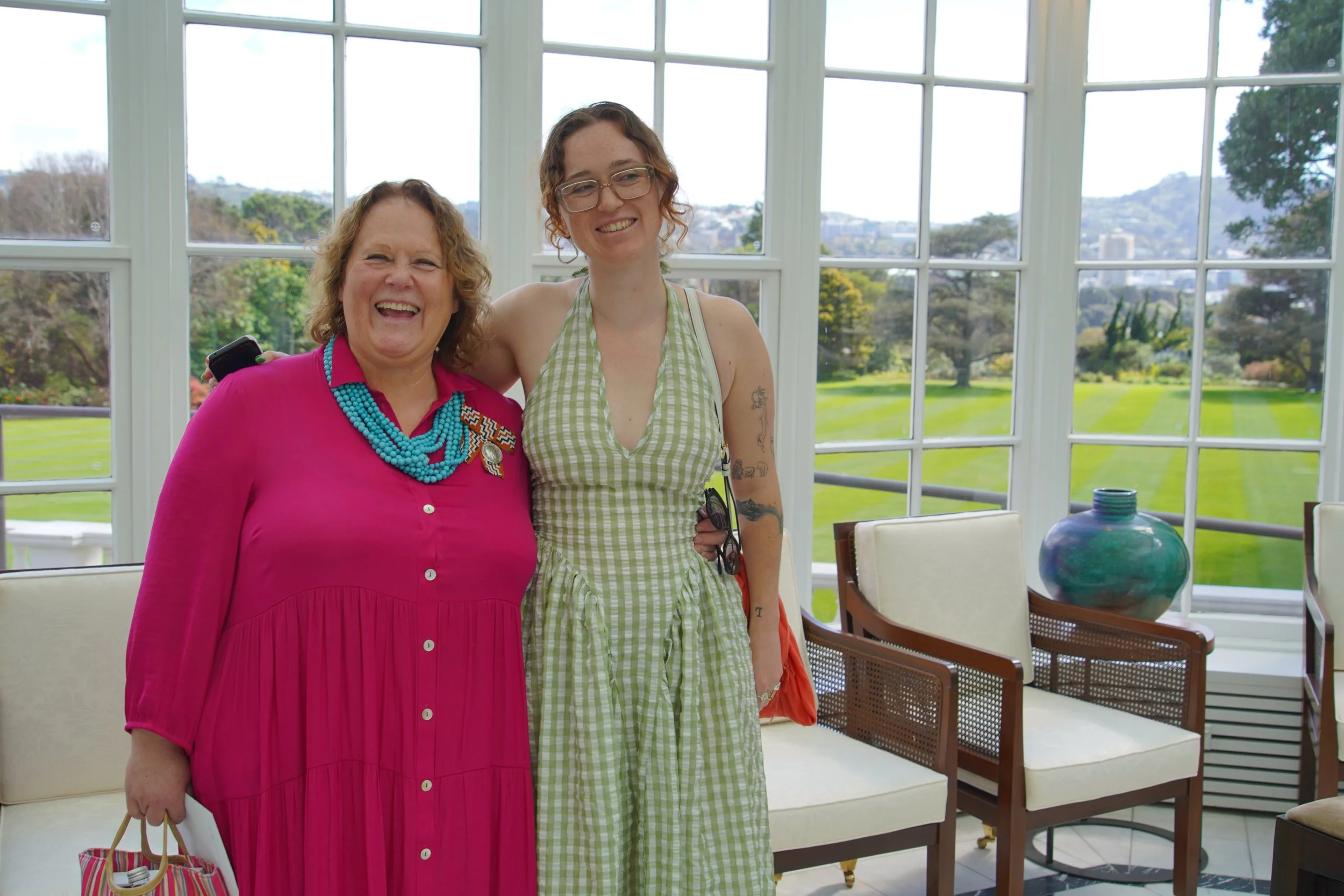 Two women standing indoors in front of large windows with a view of greenery and hills. The woman on the left has curly hair, is wearing a bright pink dress with buttons and a turquoise necklace, and is smiling broadly. The woman on the right has cur