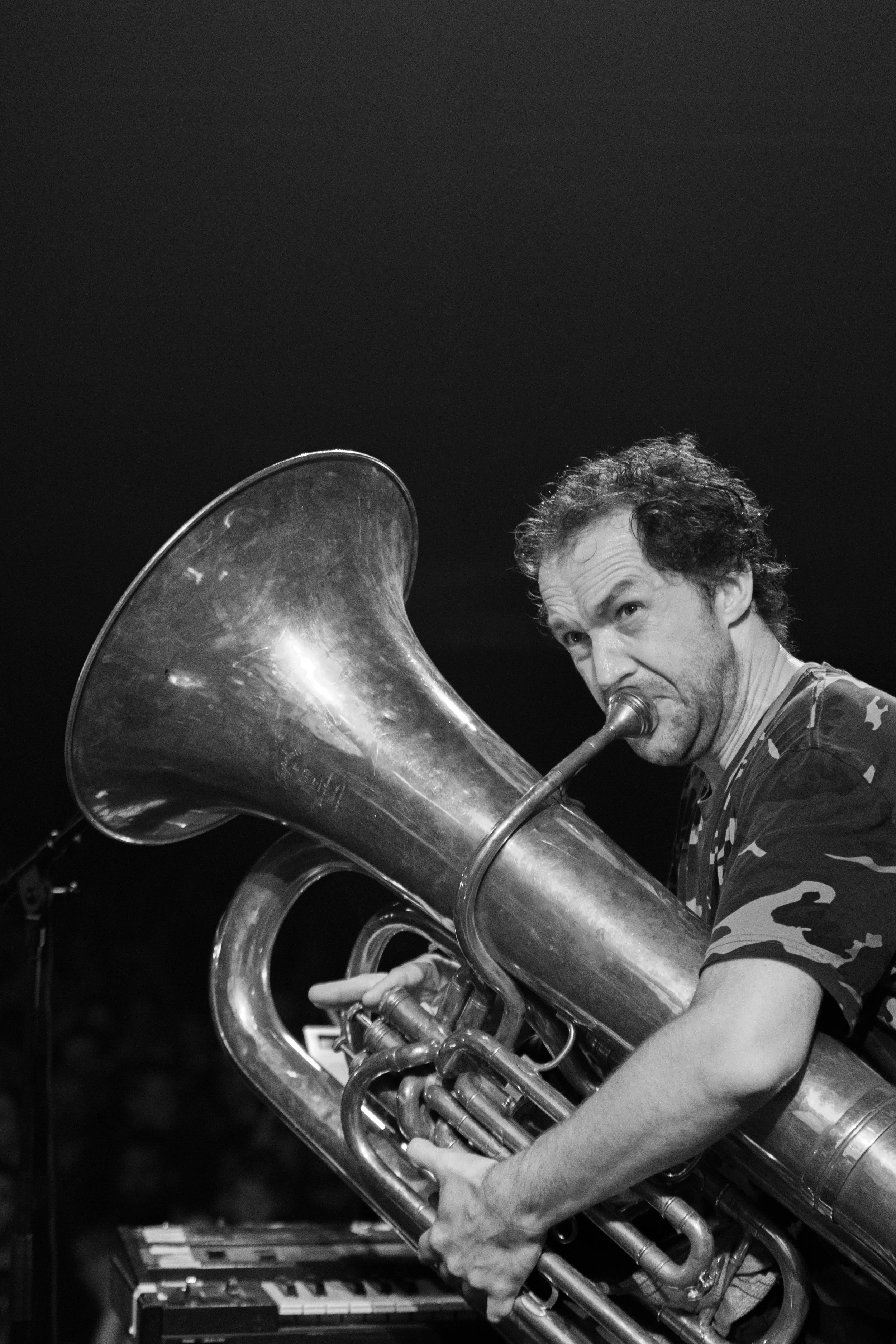 A man with curly hair playing a large brass tuba, looking at the instrument.