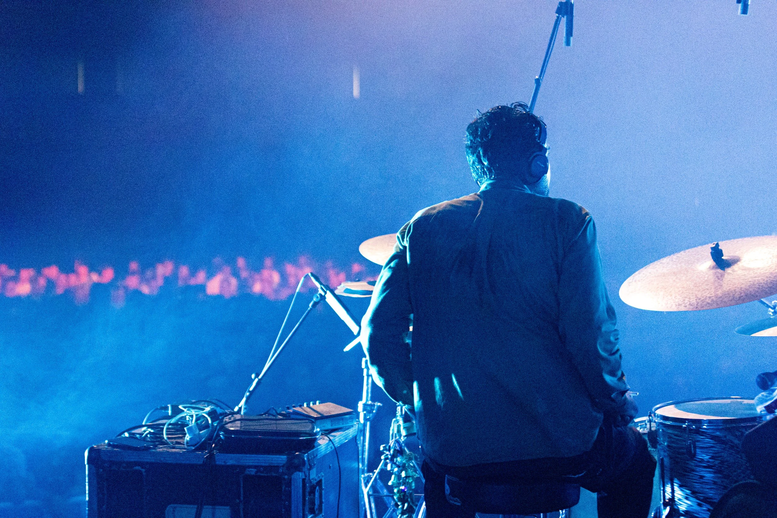 A person with dark hair, wearing headphones, is playing percussion instruments on a stage with blue lighting.