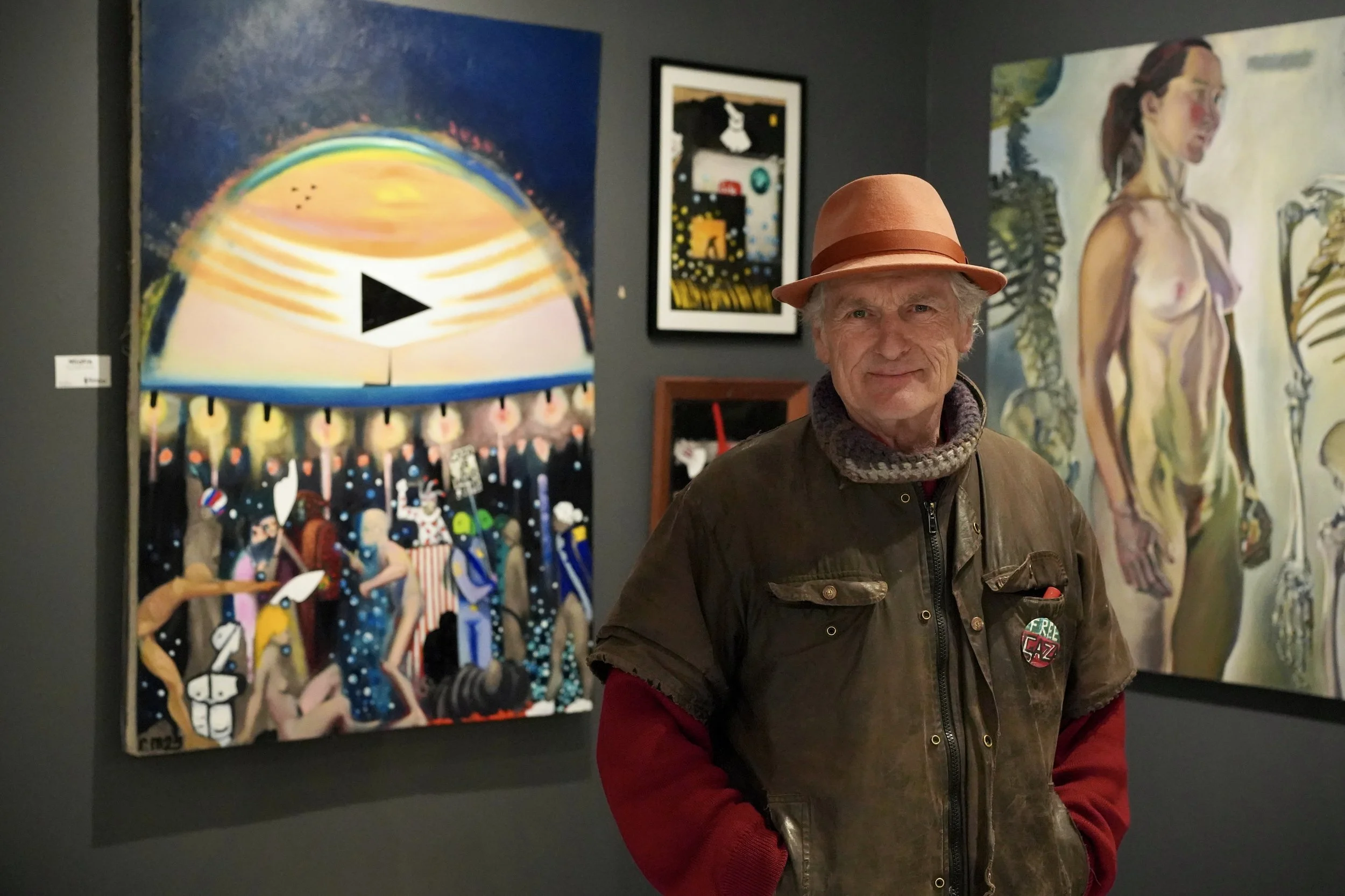 An older man with a brown jacket, scarf, and a pink hat standing in an art gallery with colorful paintings on dark walls behind him.