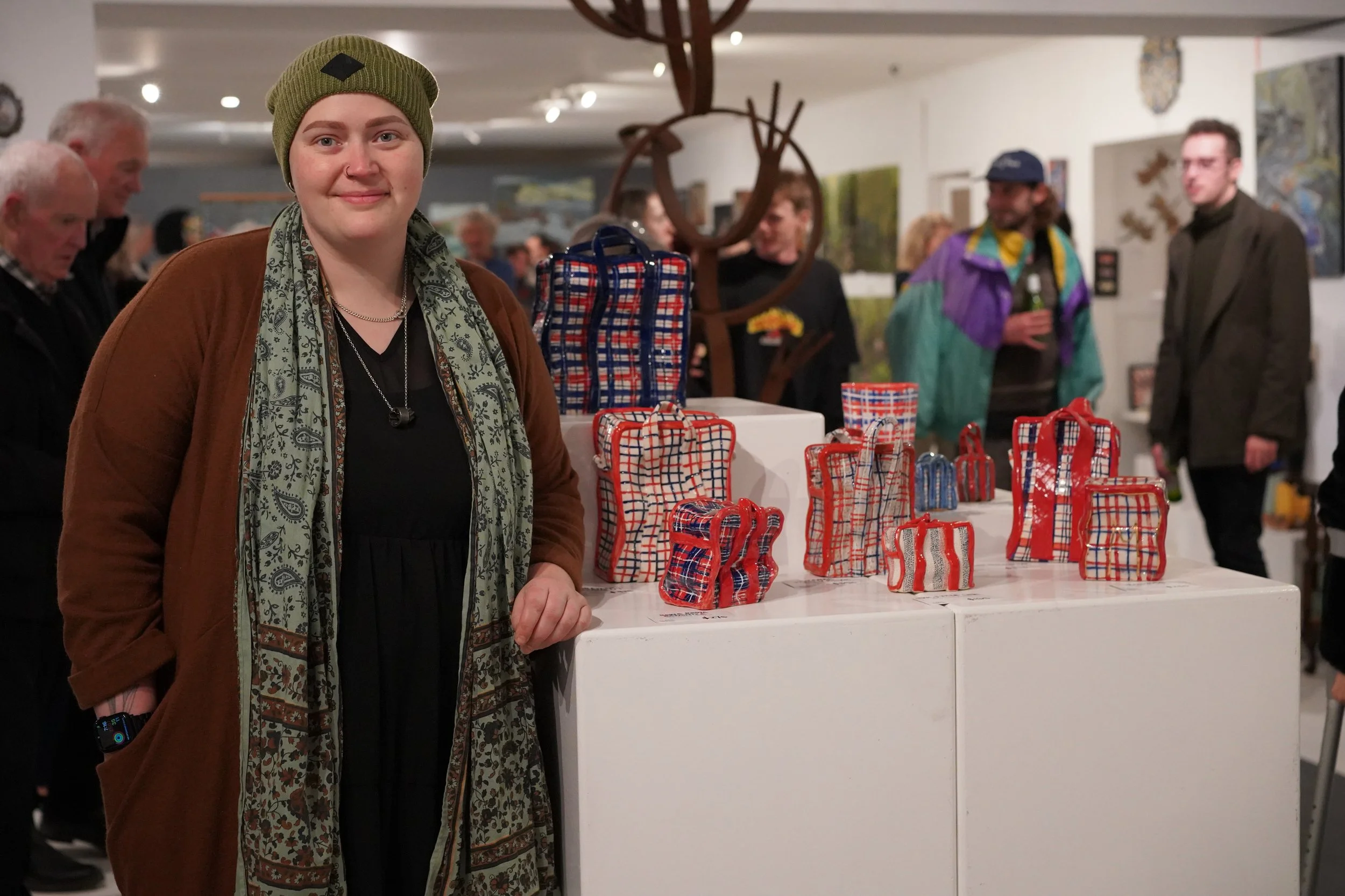 A woman with a green knit beanie, brown jacket, and patterned scarf smiling at a social event, standing near a display of red, white, and blue plaid gift bags on a white table, with people in the background at an art gallery or exhibition space.