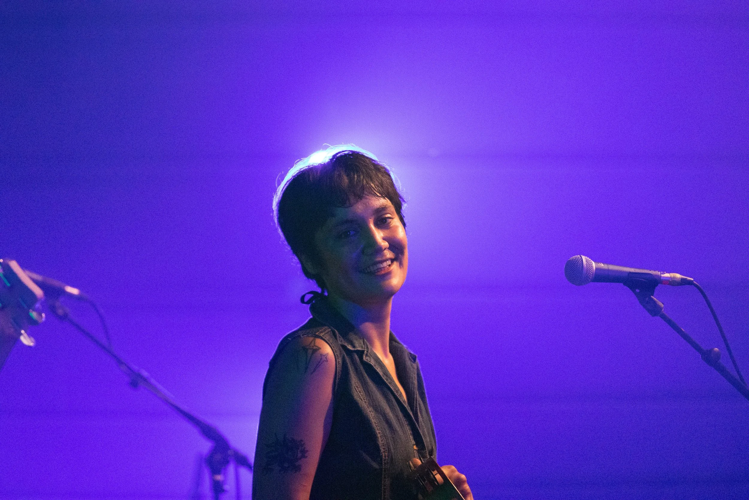 A woman with short dark hair smiling on stage with purple lighting, holding a guitar, standing near a microphone.