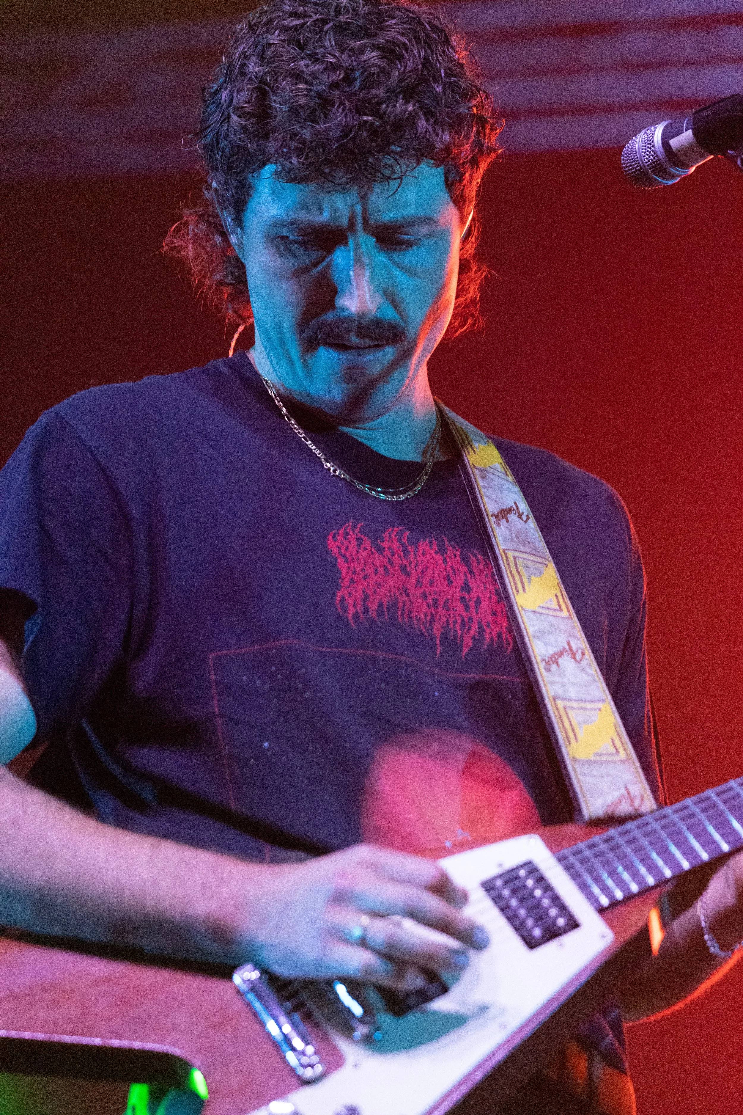 Close-up of a man with curly hair and a mustache playing an electronic keyboard, wearing a dark T-shirt with a graphic and a necklace, with a microphone nearby, in a dimly lit environment with red and purple lighting.