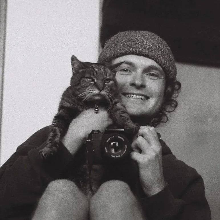 Person smiling in a mirror selfie, wearing a beanie, holding a cat close to face with one hand, taking photo with a camera in the other hand.