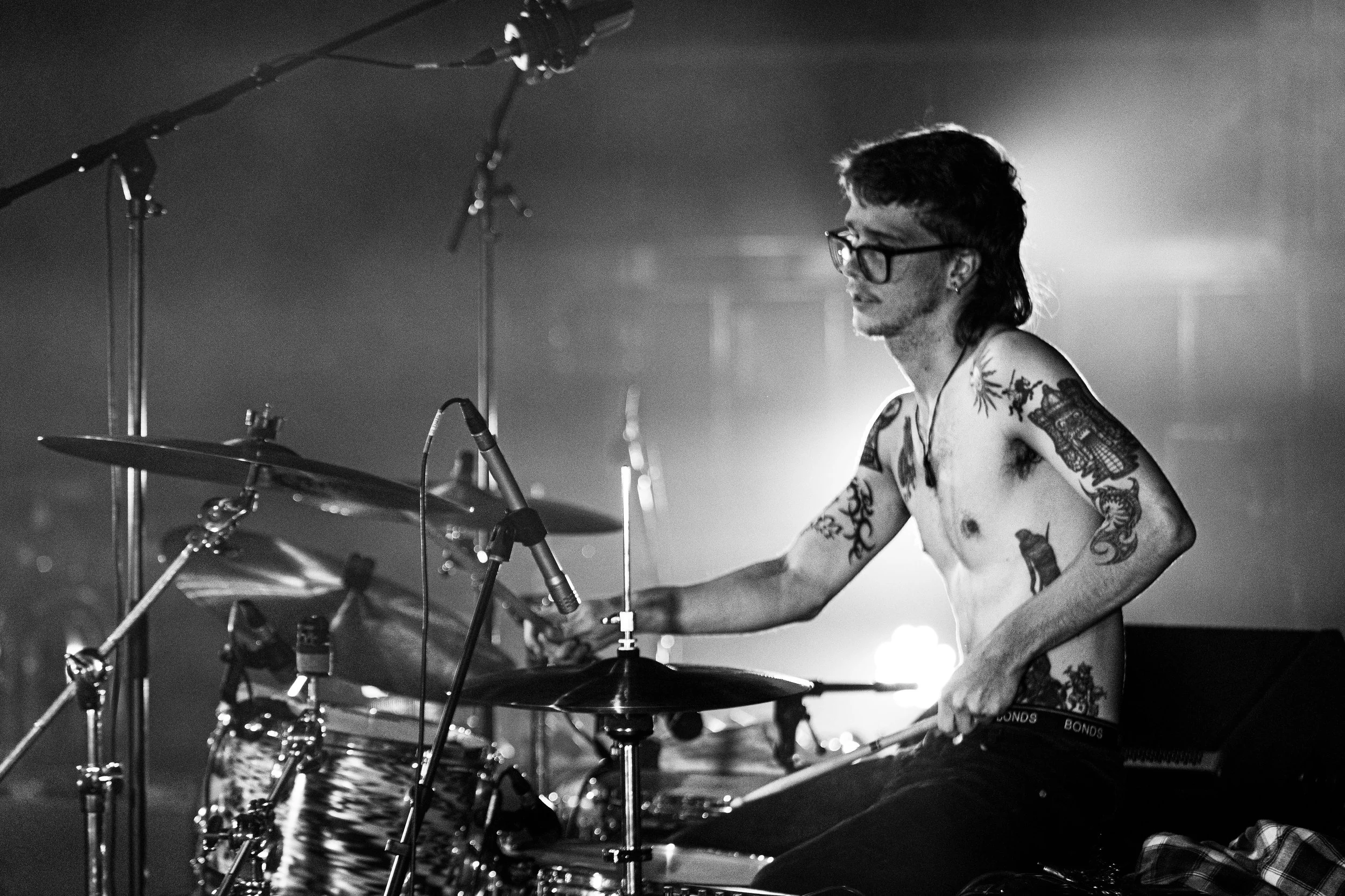 A shirtless drummer with tattoos and glasses playing drums on stage, illuminated by bright stage lights in black and white.