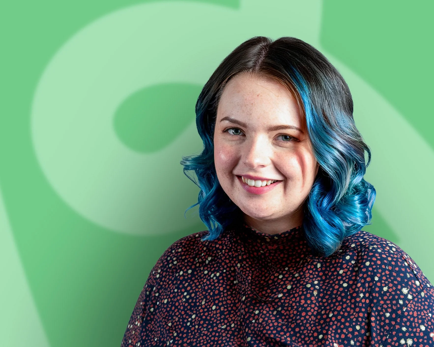 Rhian smiling at the camera with shoulder length curly blue hair, on a green background