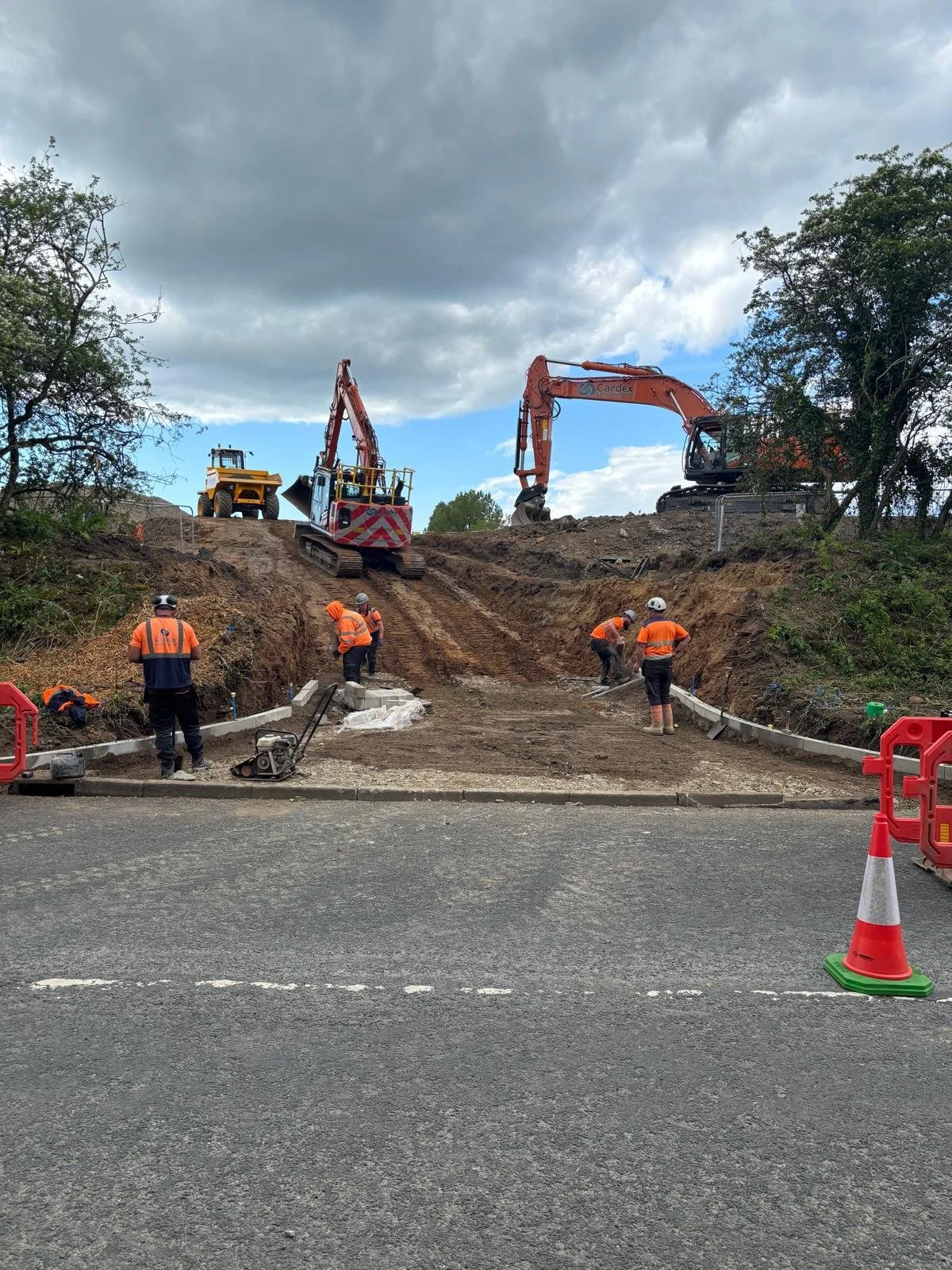 Construction workers in orange safety vests and helmets working on a road expansion project. Heavy machinery, including excavators and bulldozers, are on a dirt road with a cloudy sky overhead. Orange cones are placed on the street, indicating ongoing construction.
