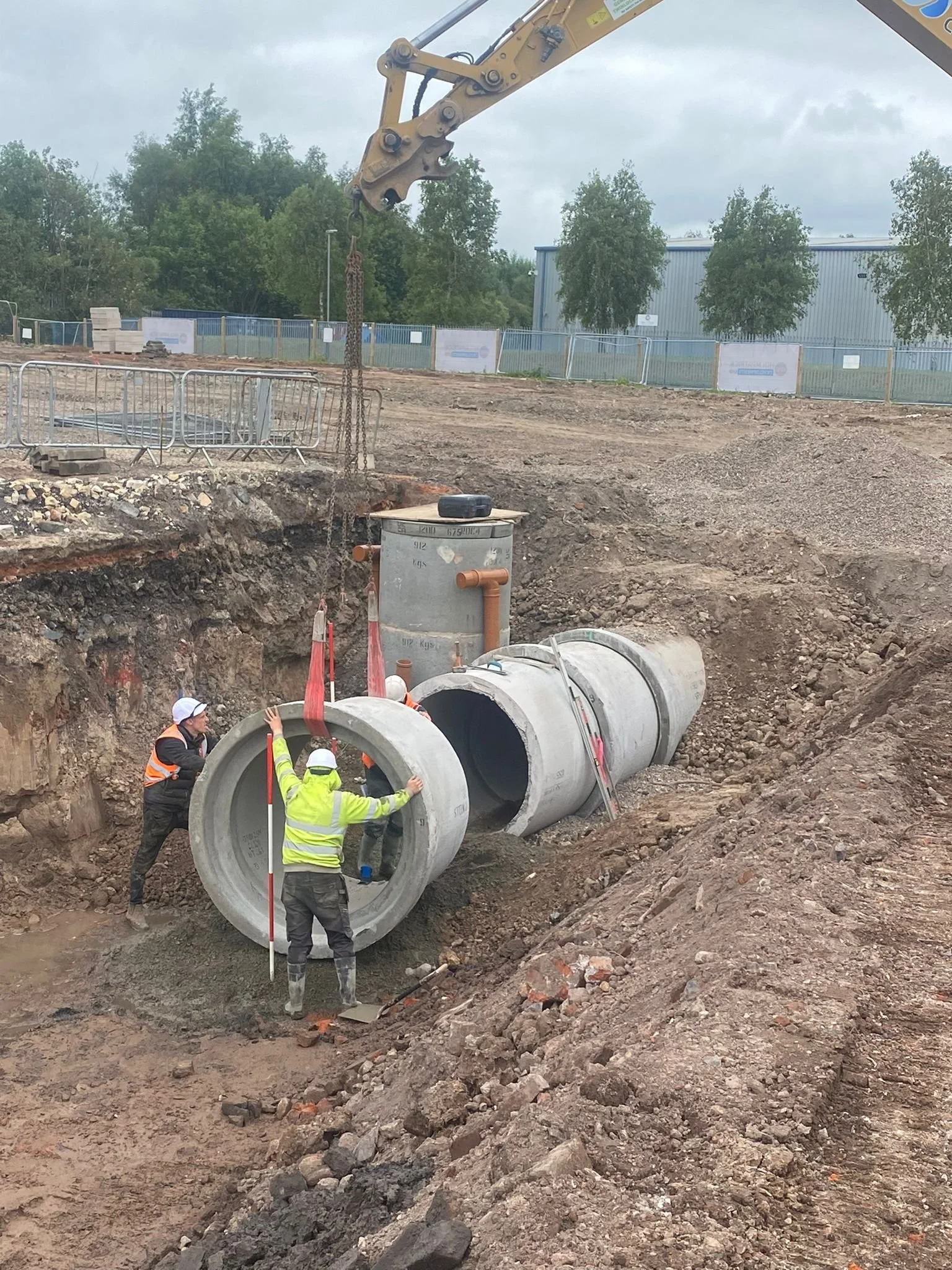Construction workers installing large concrete pipes into a large trench at a construction site. A crane lifts a pipe into place, and workers guide the pipe into position.
