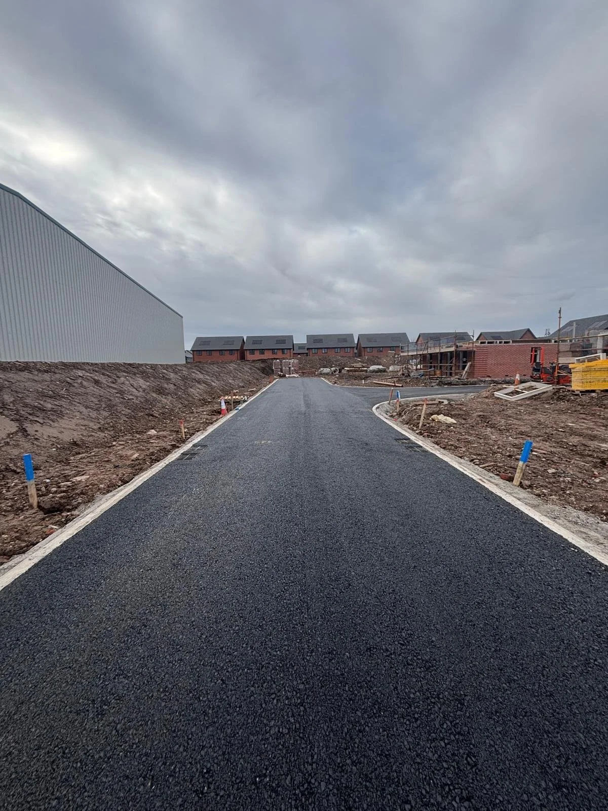 Newly paved asphalt road under construction in a residential area with houses and construction materials in the background and cloudy sky.