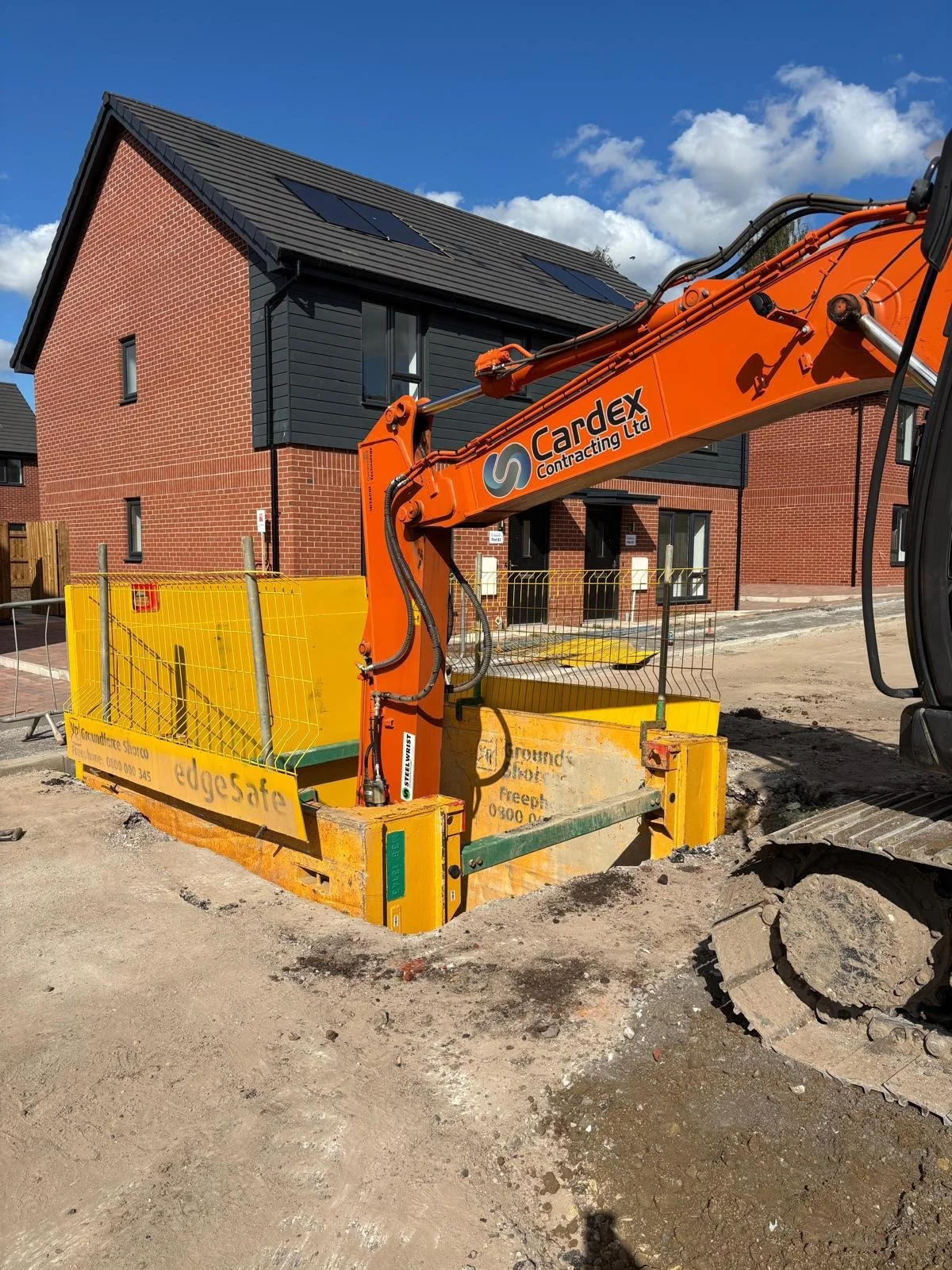 Construction site with an orange excavator lifting a yellow barrier in front of a new brick residential building with black accents and solar panels on the roof, under a blue sky with scattered clouds.
