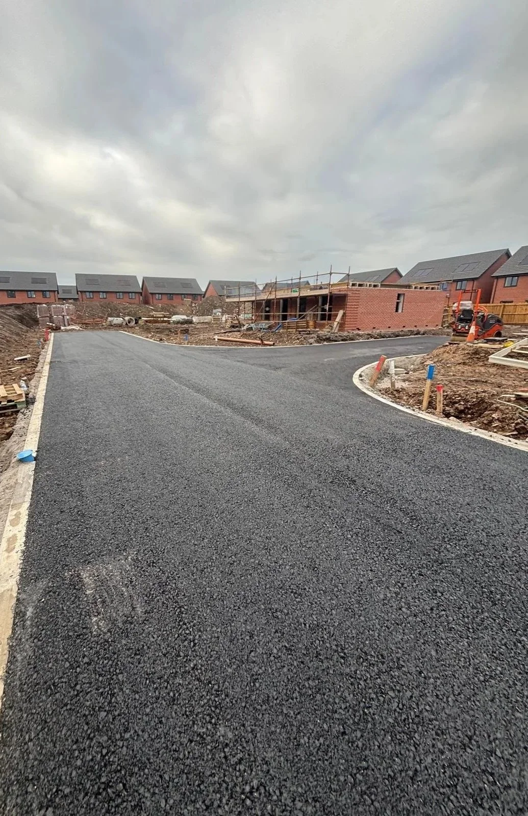 A newly paved asphalt road under construction in a residential area, with houses in the background and construction equipment around.