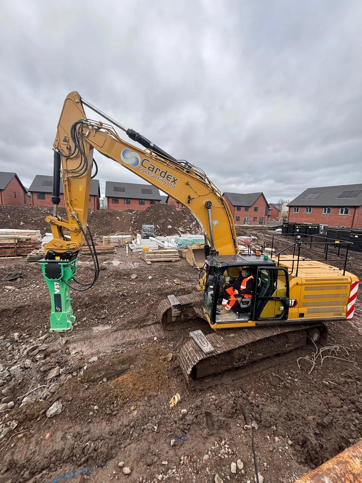 A construction excavator with a worker in the cab at a building site with houses in the background, cloudy sky.