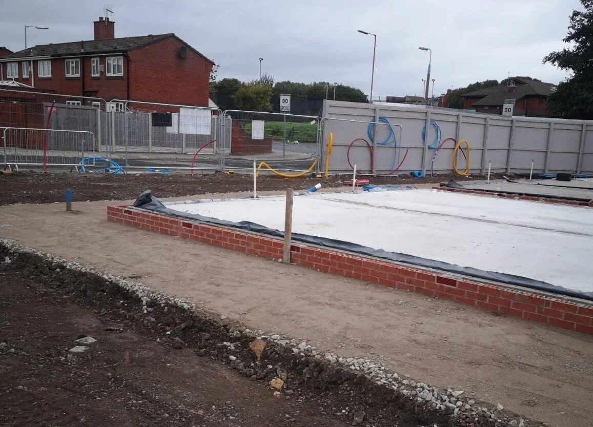 Construction site with a brick outline for a building foundation, surrounded by construction barriers and plumbing pipes, in a residential area.