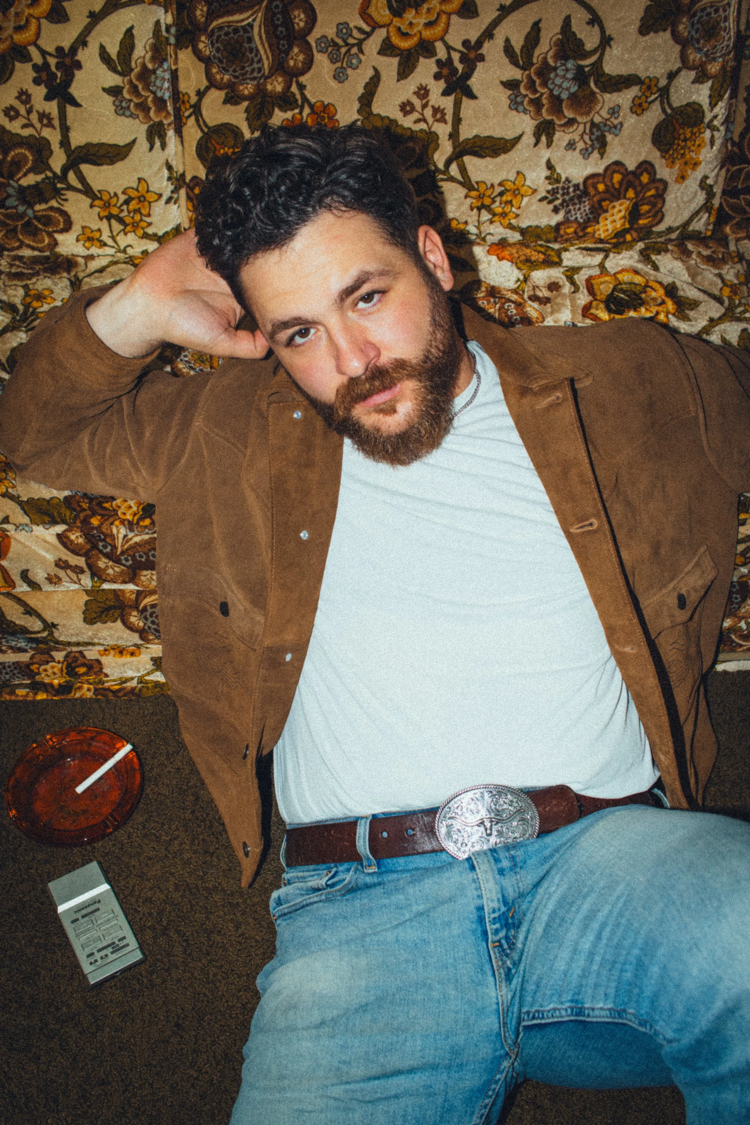 A man with dark curly hair and a beard lying on a floral patterned sofa, wearing a white t-shirt, brown jacket, and blue jeans with a large belt buckle. There is a drink in a red glass and a remote control on the floor nearby.