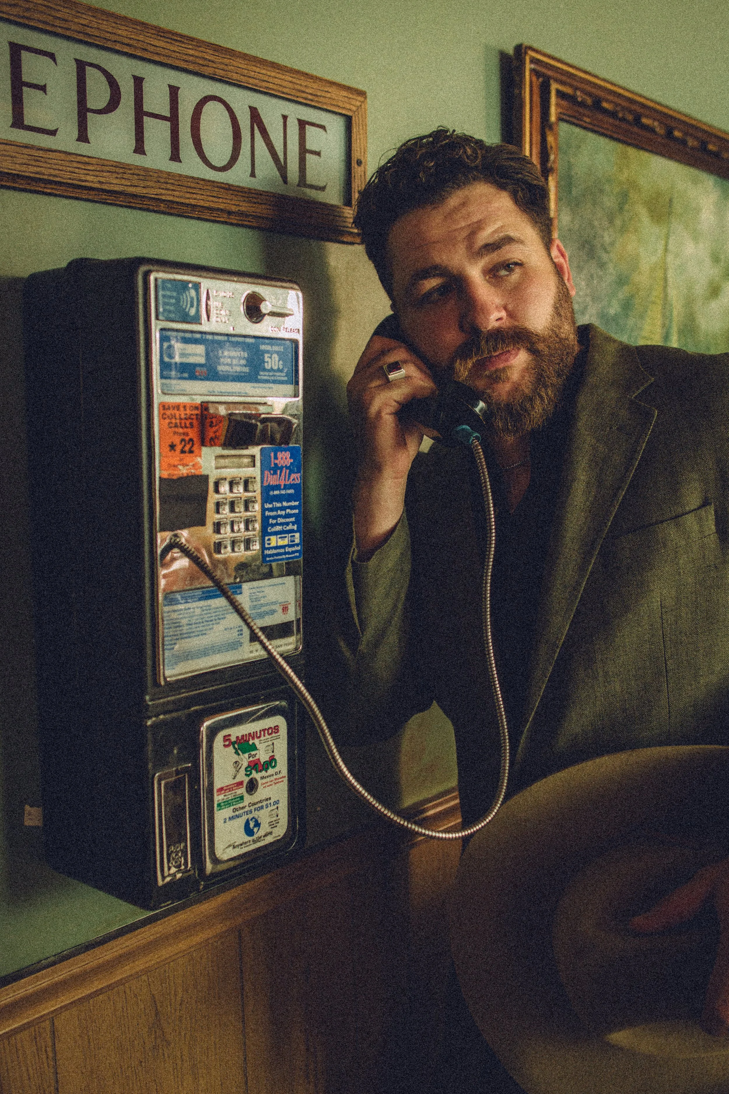 A man with a beard and mustache talking on a payphone in a dimly lit room, with a wall map and framed picture in the background.
