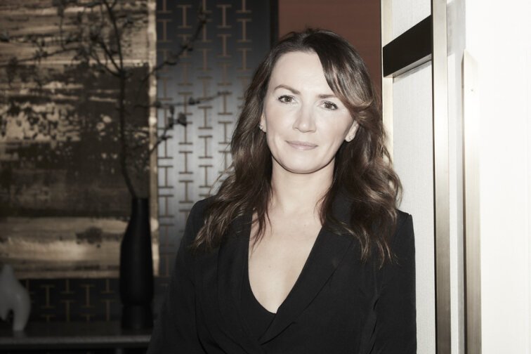 A woman with wavy brown hair and light makeup posing indoors near a window and decorative wall pattern.